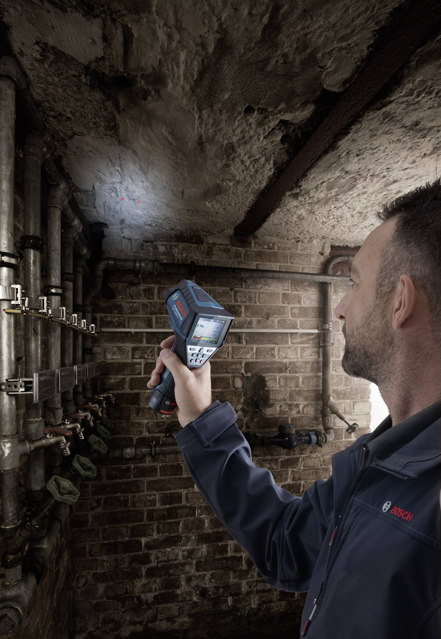 Een man gebruikt een handapparaat om de vochtigheid te meten aan een kelderplafond. Op de achtergrond zijn buizen en een bakstenen muur zichtbaar.