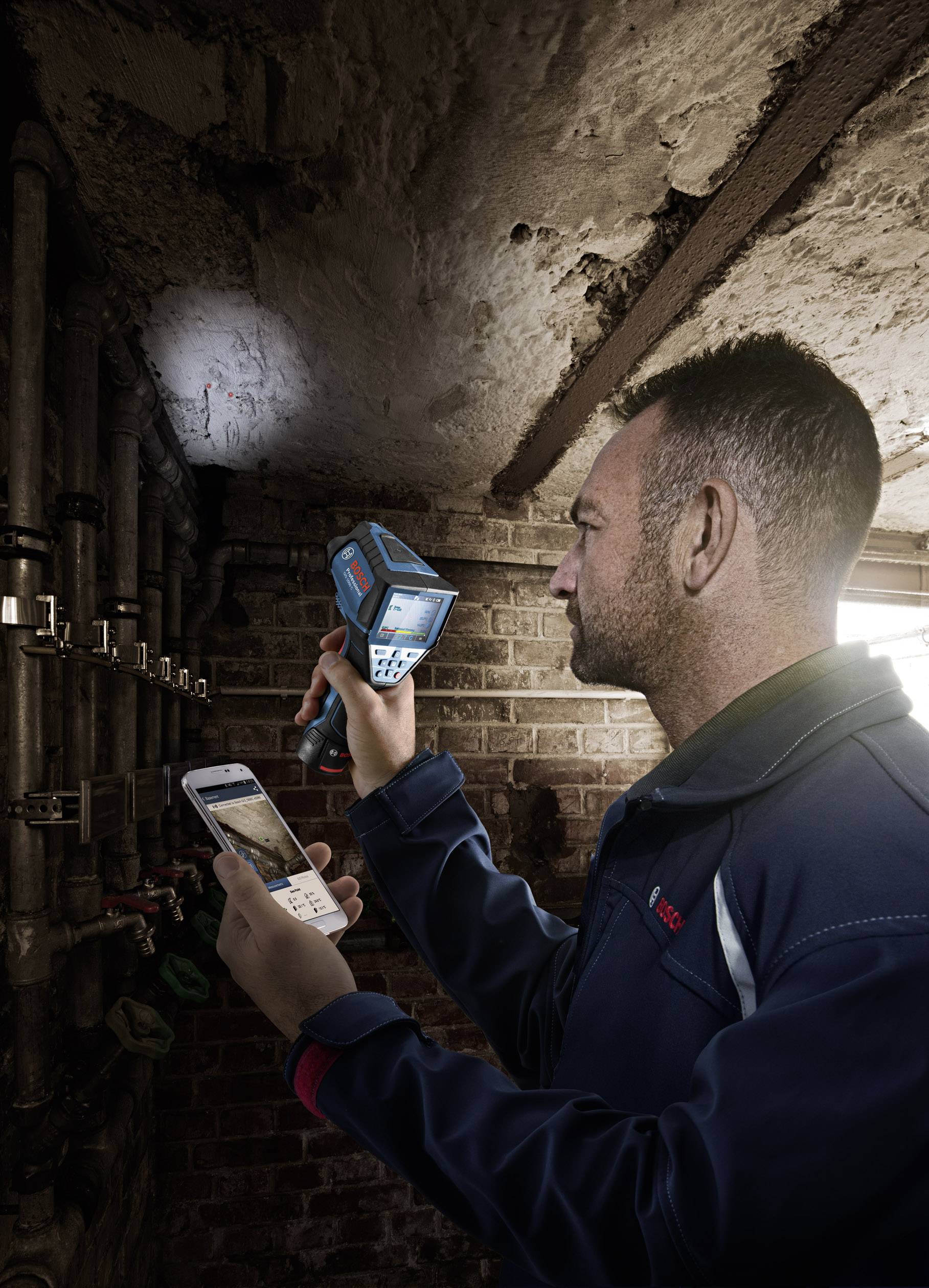 Een man inspecteert een pijpleiding in een kelder met een warmtebeeldcamera en een smartphone om de temperatuur te controleren.