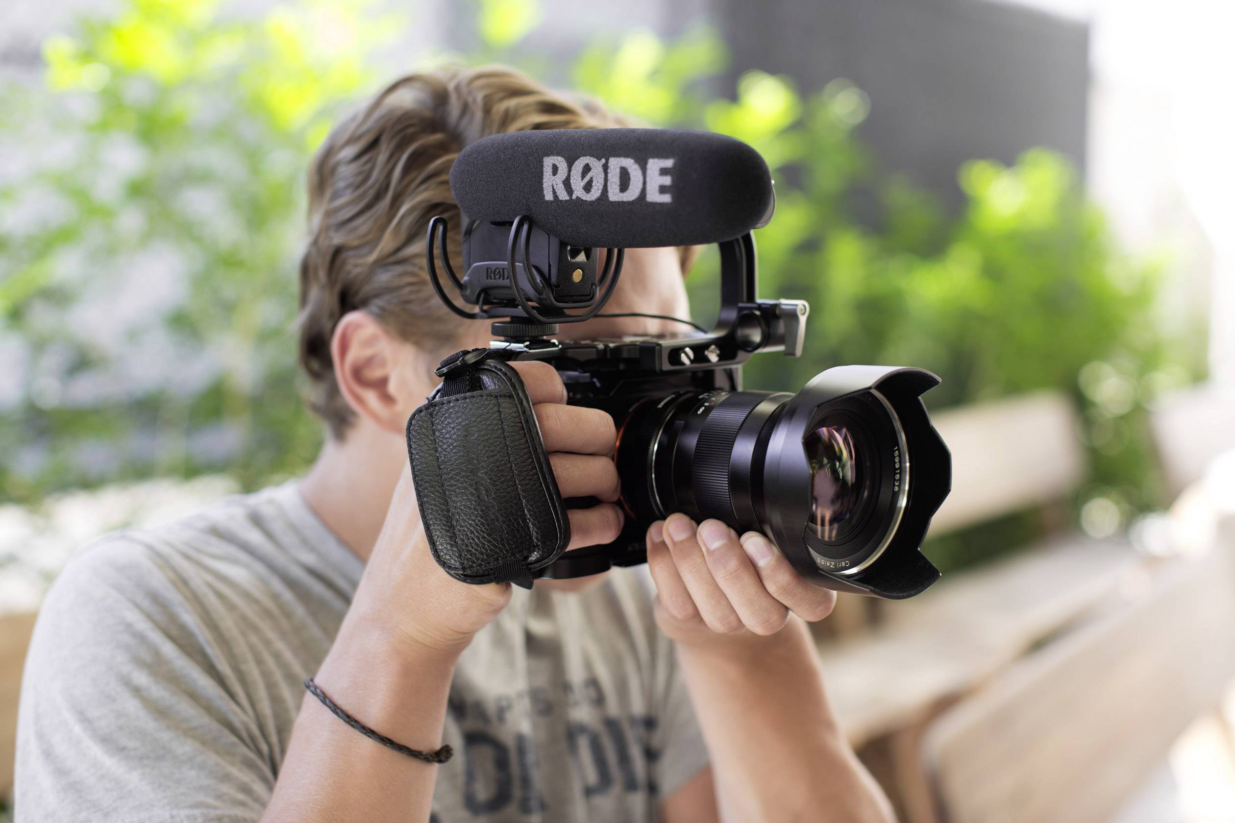 Een persoon filmt met een camera die is uitgerust met een RØDE-microfoon. De achtergrond toont groene planten in de buitenlucht.