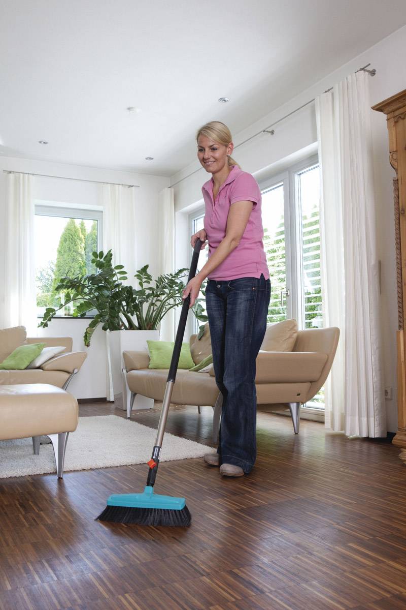 Persoon in roze shirt dweilt houten vloer in gezellige woonkamer met planten en banken. Lichtinval door grote ramen.