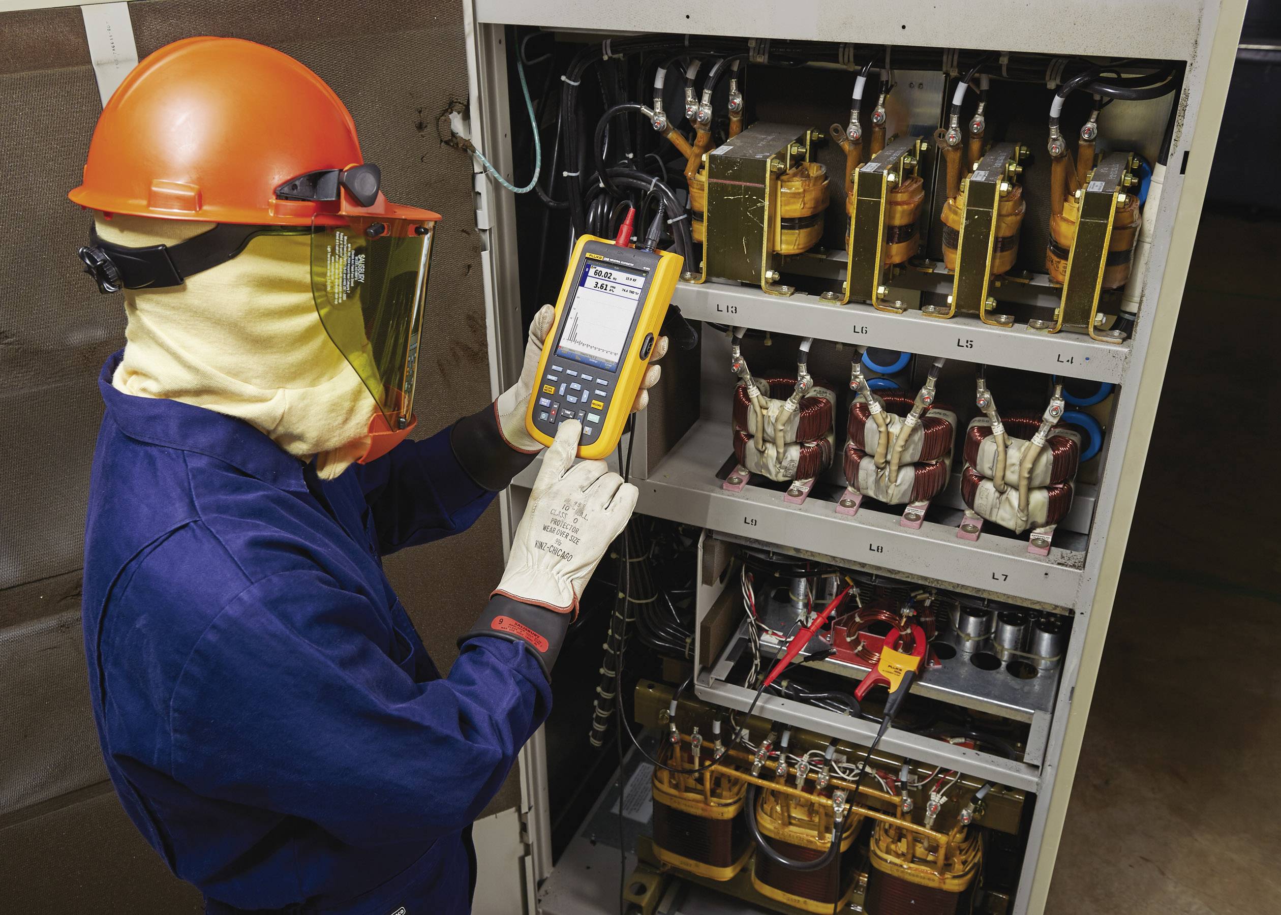 Een technicus in beschermende uitrusting meet met een draagbaar apparaat de stroom in een open schakelkast met transformatoren.