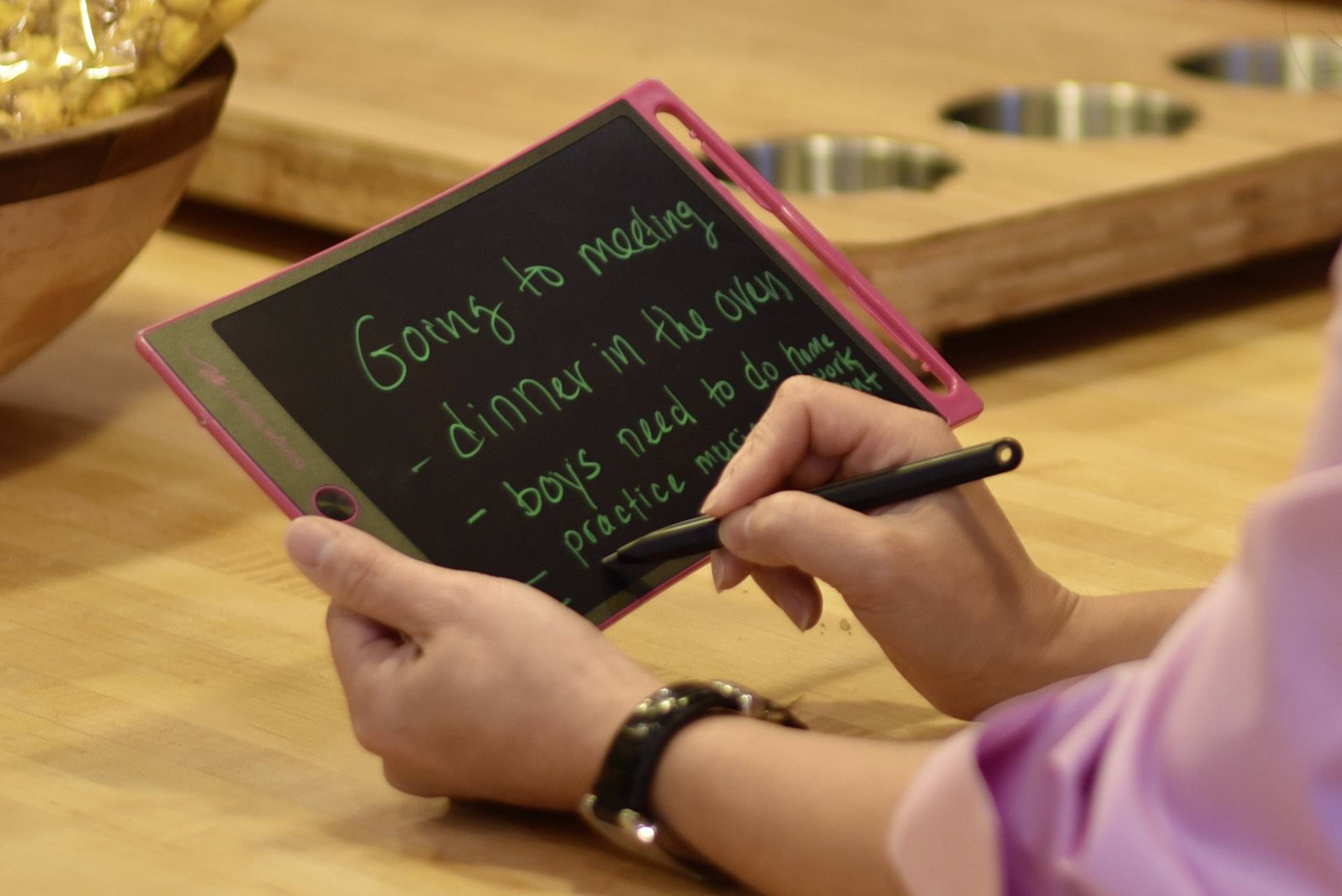 Een persoon houdt een elektronisch schrijftablet vast. Op het display staat: 'Naar vergadering, eten in de oven, jongens moeten huiswerk maken, muziek oefenen'.
