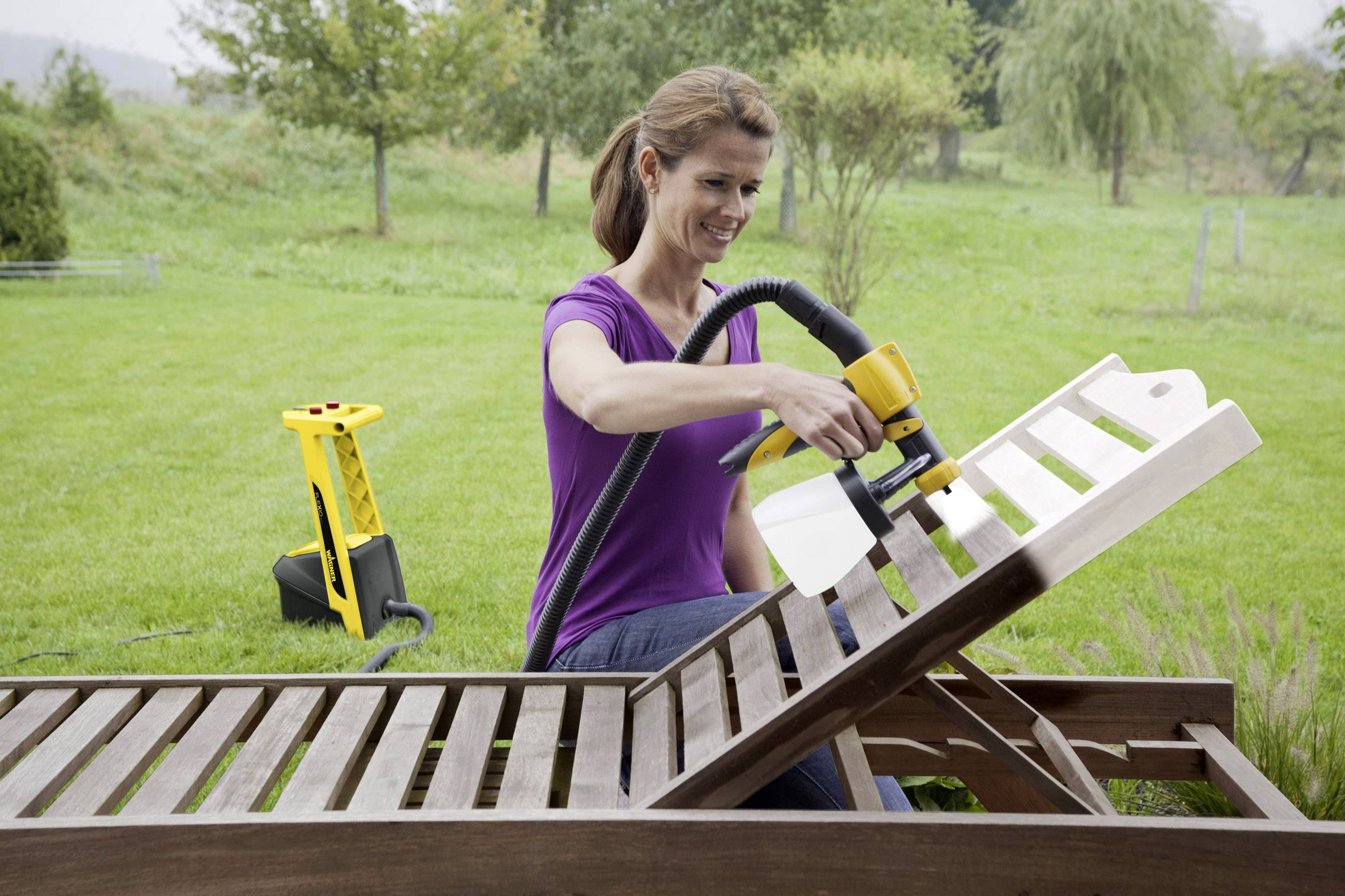 Een vrouw beschildert een houten ligstoel met een verfspuitbus in de tuin. Op de achtergrond zijn bomen en een groene weide zichtbaar.
