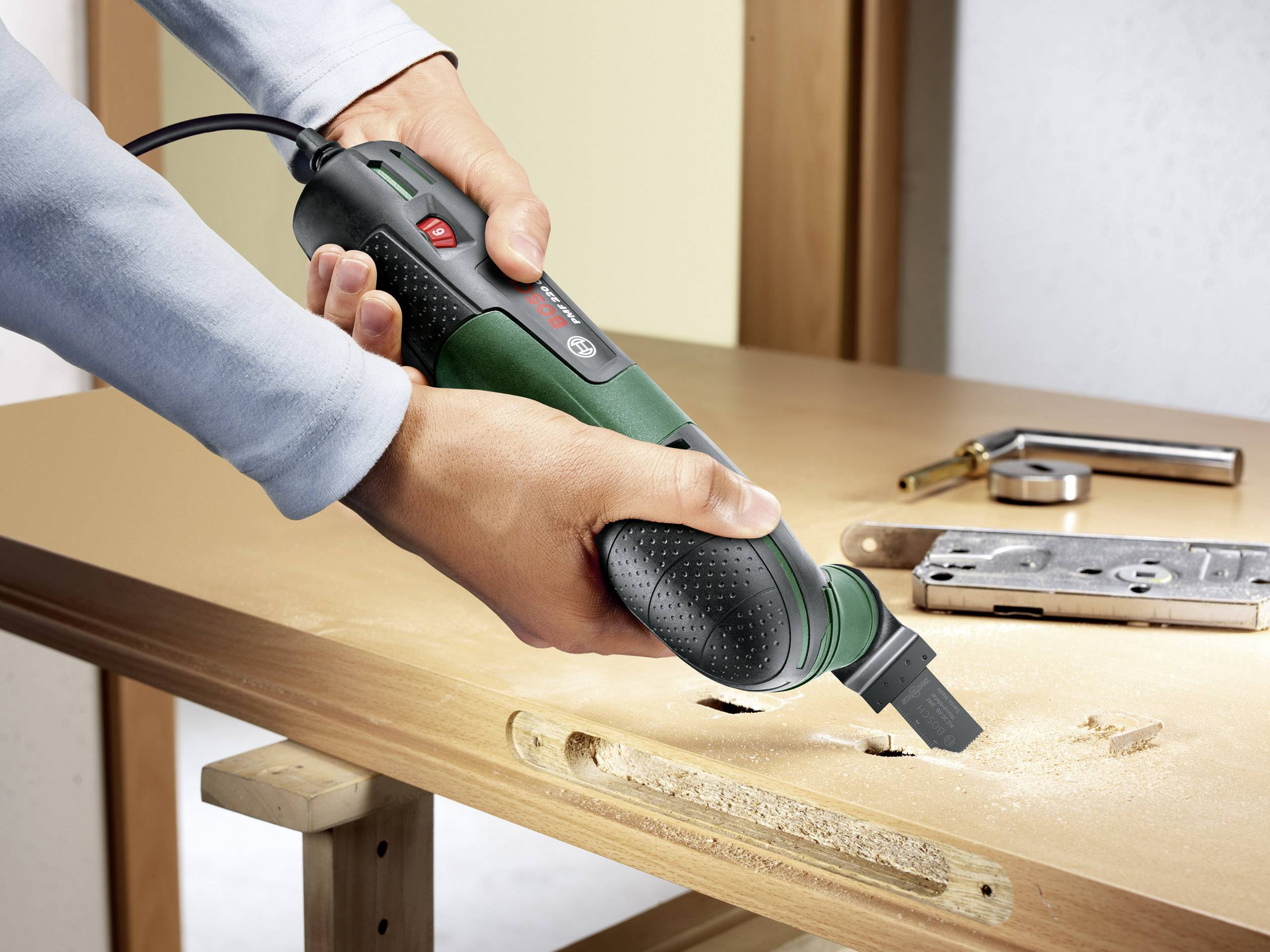 Een persoon gebruikt een elektrisch multifunctioneel gereedschap om een rechthoekig gat uit te zagen in een houten tafel. Het gereedschap heeft een groen behuizing.