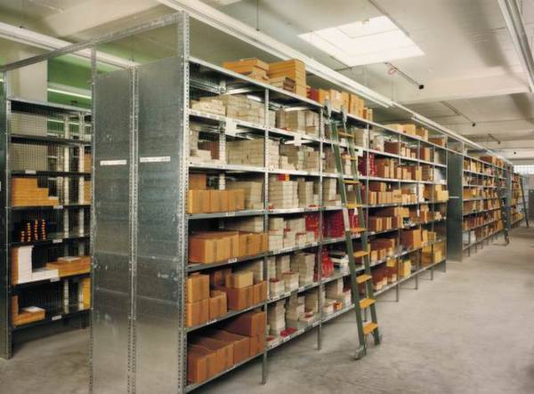 Opslagruimte met metalen rekken, gevuld met dozen en ordners; een ladder staat aan de zijkant voor toegang tot de hogere schappen.