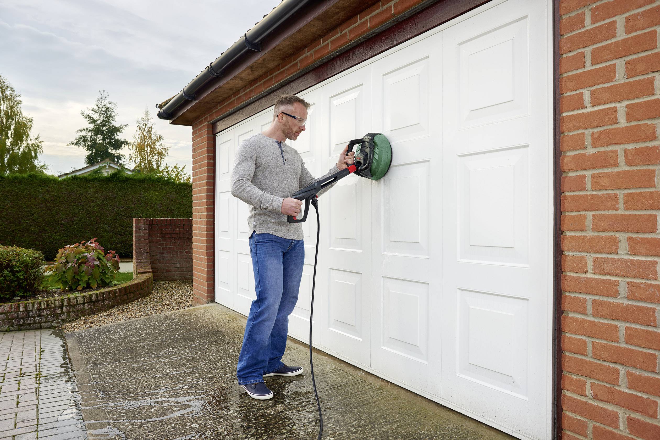 Een man in spijkerbroek en trui reinigt een wit garagedeur met een hogedrukreiniger aan een bakstenen gebouw.