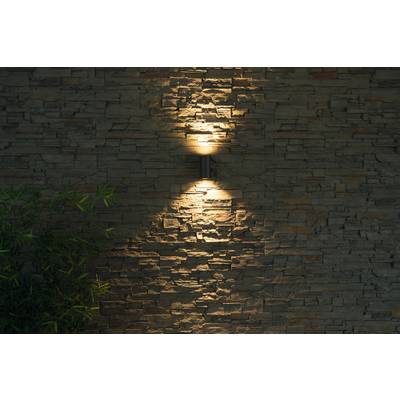 Muur van natuursteen, verlicht door een lamp die warm licht naar boven en beneden uitstraalt; linksonder zijn bamboeplanten zichtbaar.