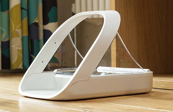 Witte automatische voederdispenser voor huisdieren op een houten vloer, geplaatst voor een gordijn en een houten meubelstuk.