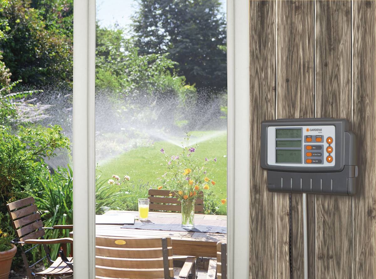 Een irrigatiebesturingsapparaat hangt aan een houten muur met uitzicht op een tuin. In de tuin wordt water gespoten, en buiten staat een tafel.