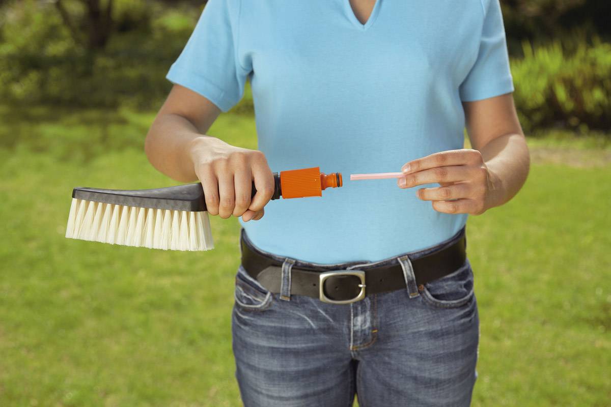 Een persoon houdt een schoonmaakborstel vast en steekt een lange roze stang door de handgreep, terwijl er gras te zien is op de achtergrond.