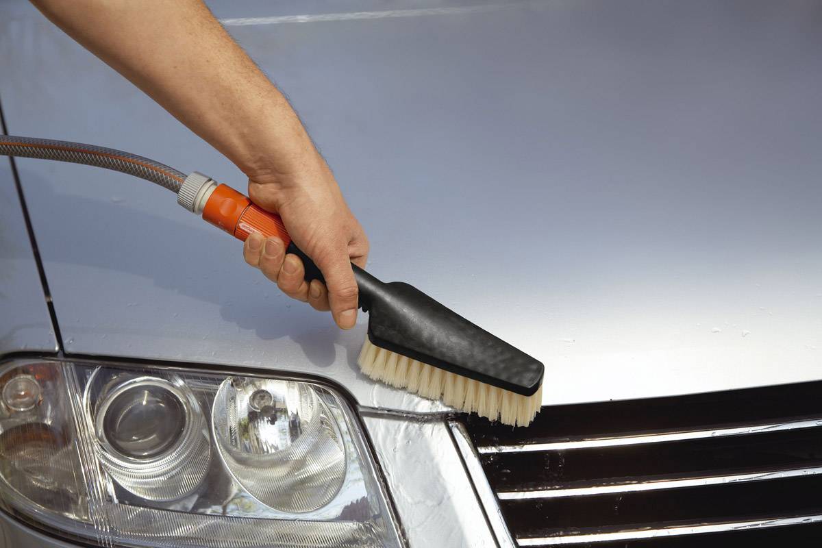 Hand met borstel reinigt een zilveren auto-radiateurrooster, gefocust op de beweging van de borstel over het oppervlak nabij de koplamp.