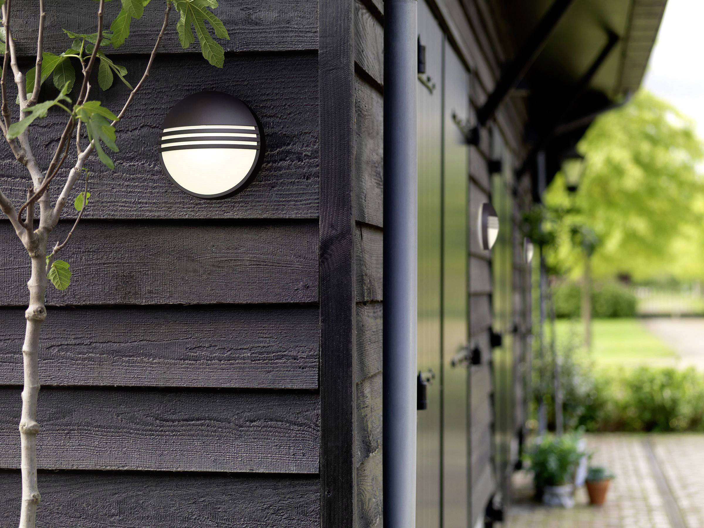 Een buitenmuurlamp schijnt aan een zwarte houten gevel naast een deur. Op de voorgrond staat een kleine boom; planten zijn zichtbaar.