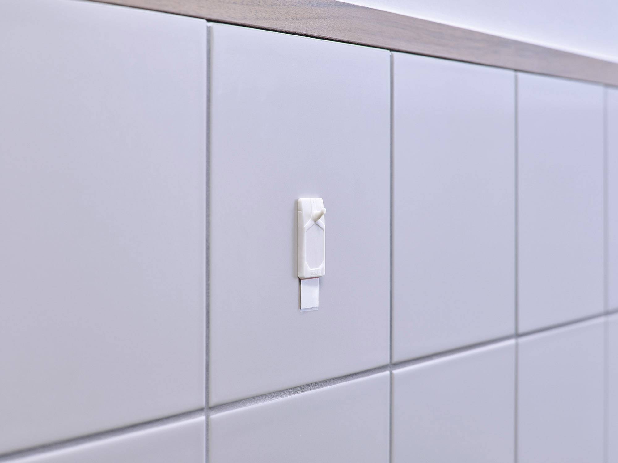 Een witte kunststof houder is bevestigd aan een betegelde muur. De houder heeft een verwijderbare of los lijkende band.