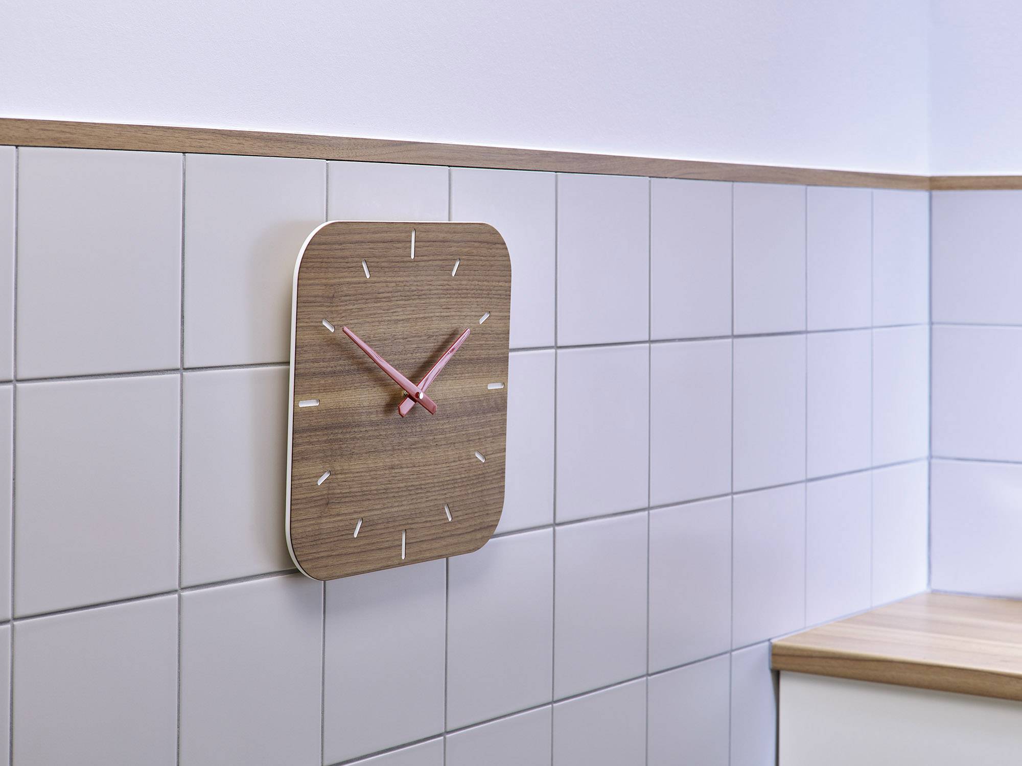 Een moderne wandklok met houtlook en roze wijzers hangt aan een wit betegelde muur in een keuken.