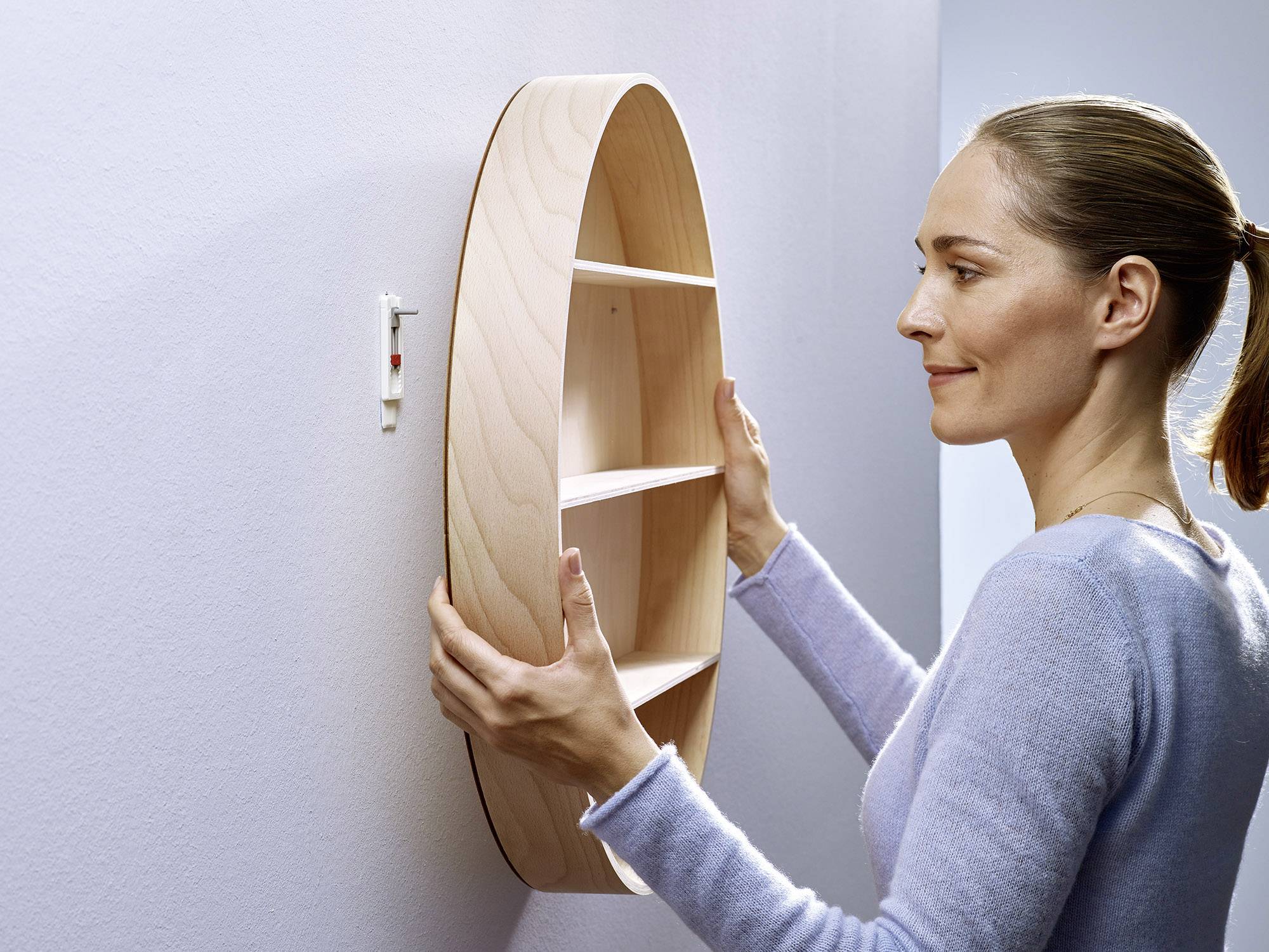 Een persoon hangt een rond, houten rek met meerdere vakken aan een witte muur.