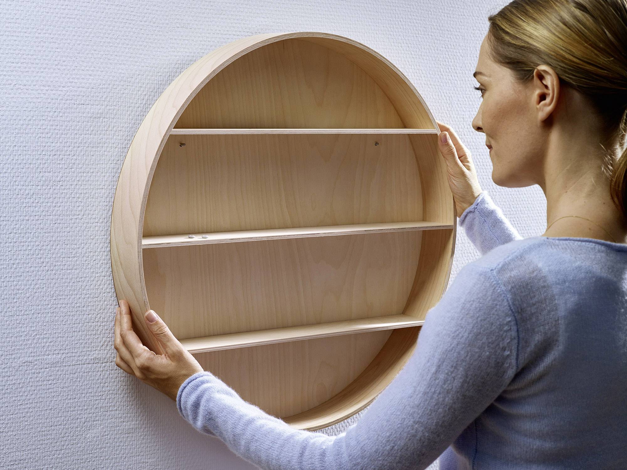 Een vrouw hangt een rond houten rek aan de muur. Het rek heeft drie planken en is bedoeld voor decoratie of opslag.