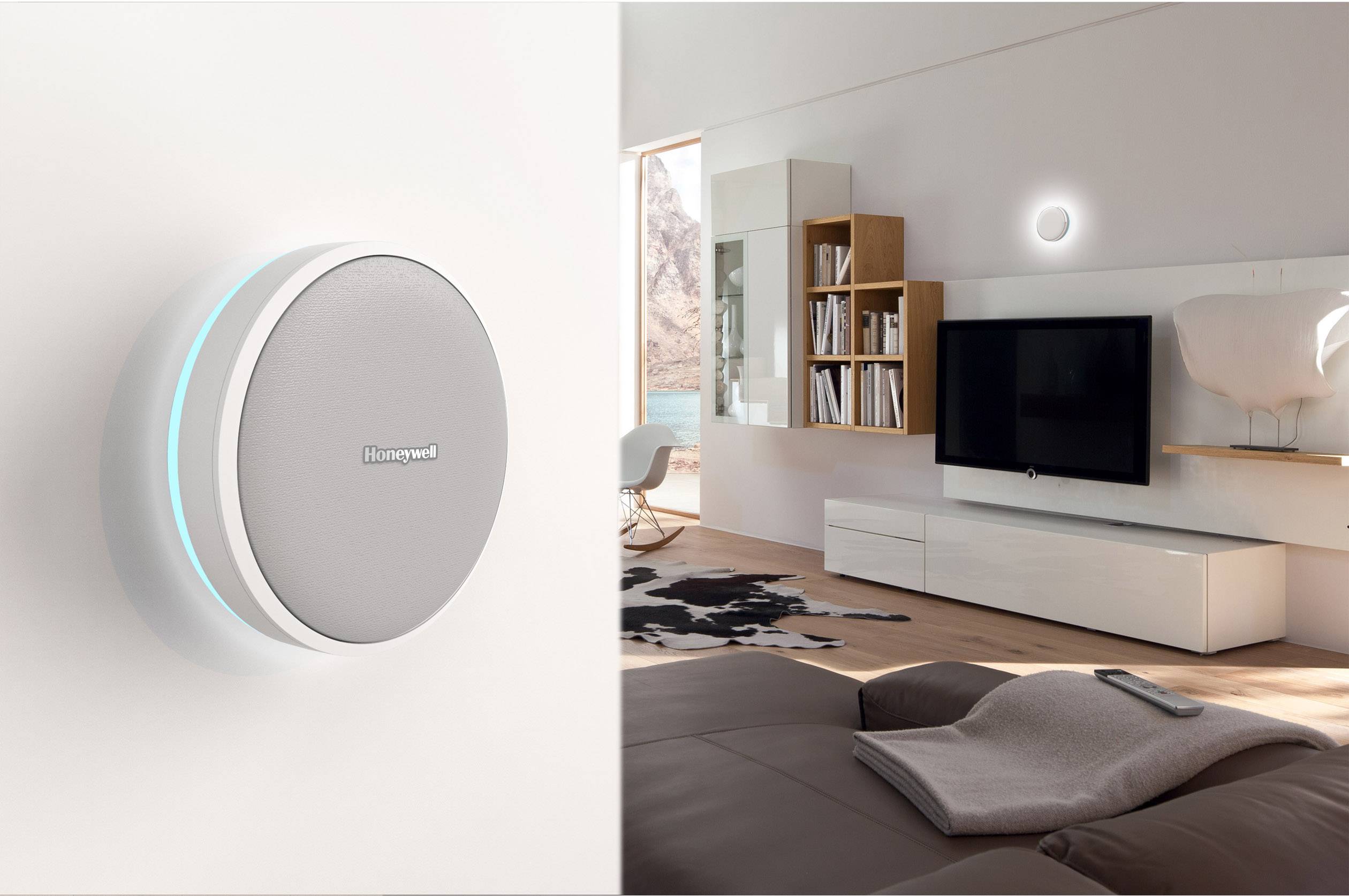 Een modern woonkamer met een grijze bank, televisie en planken. Aan de linkermuur een rond, zilver smart home-apparaat.