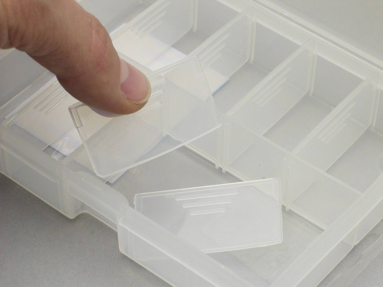 Een hand haalt een lege objectdrager uit een plastic doos met meerdere vakken.