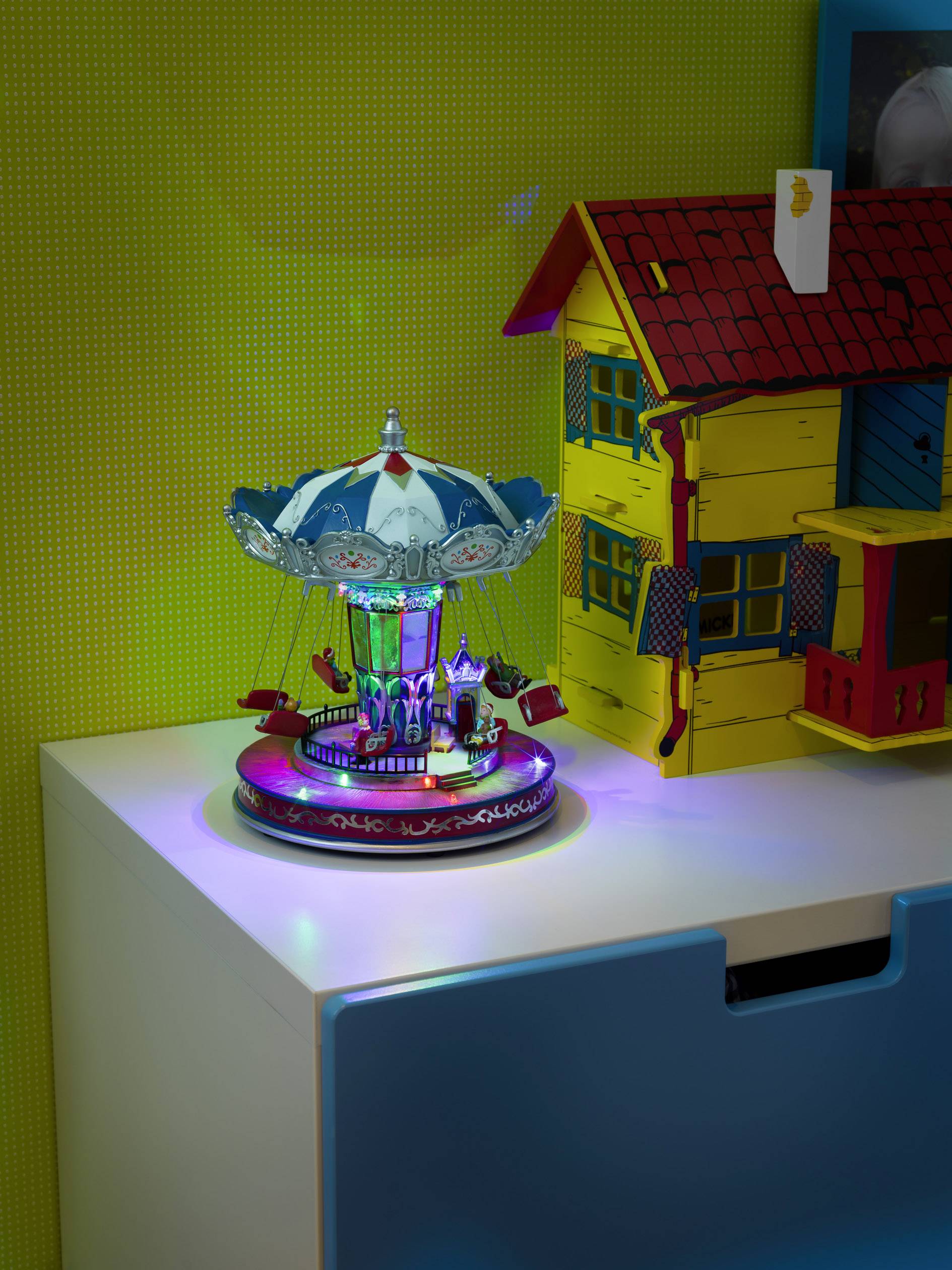 Een kleurrijk, verlicht carrouselspeelgoed staat op een dressoir naast een modelhuis. De achtergrond is geel.