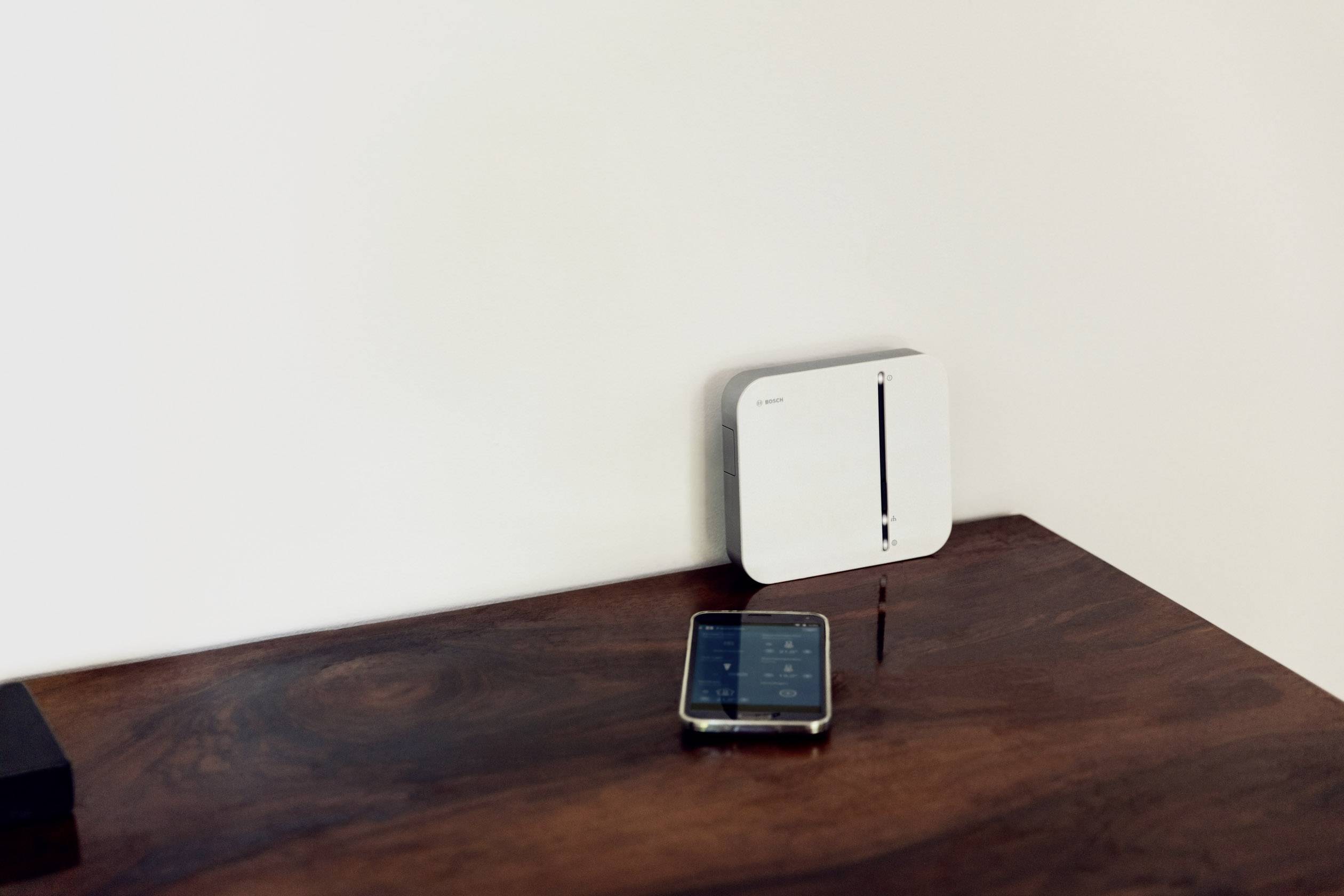 Een smartphone ligt op een houten tafel naast een wit elektronisch apparaat aan de muur.