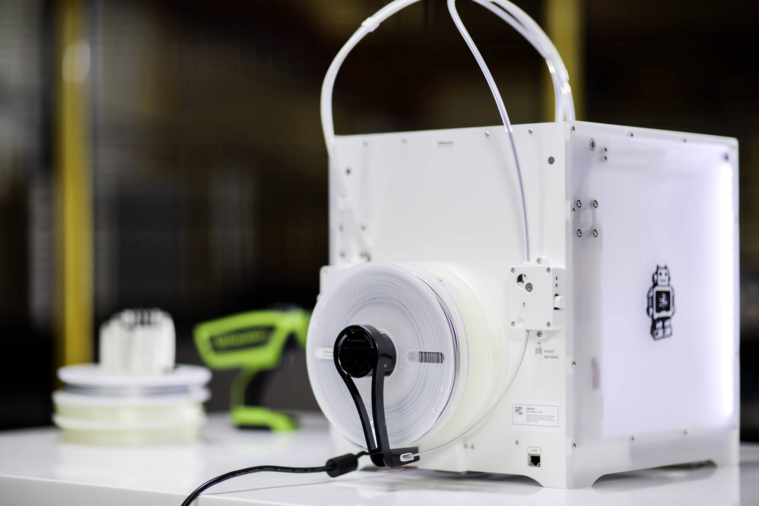 Een 3D-printer met een filamentrol, op de achtergrond meer rollen en een groen gereedschap.