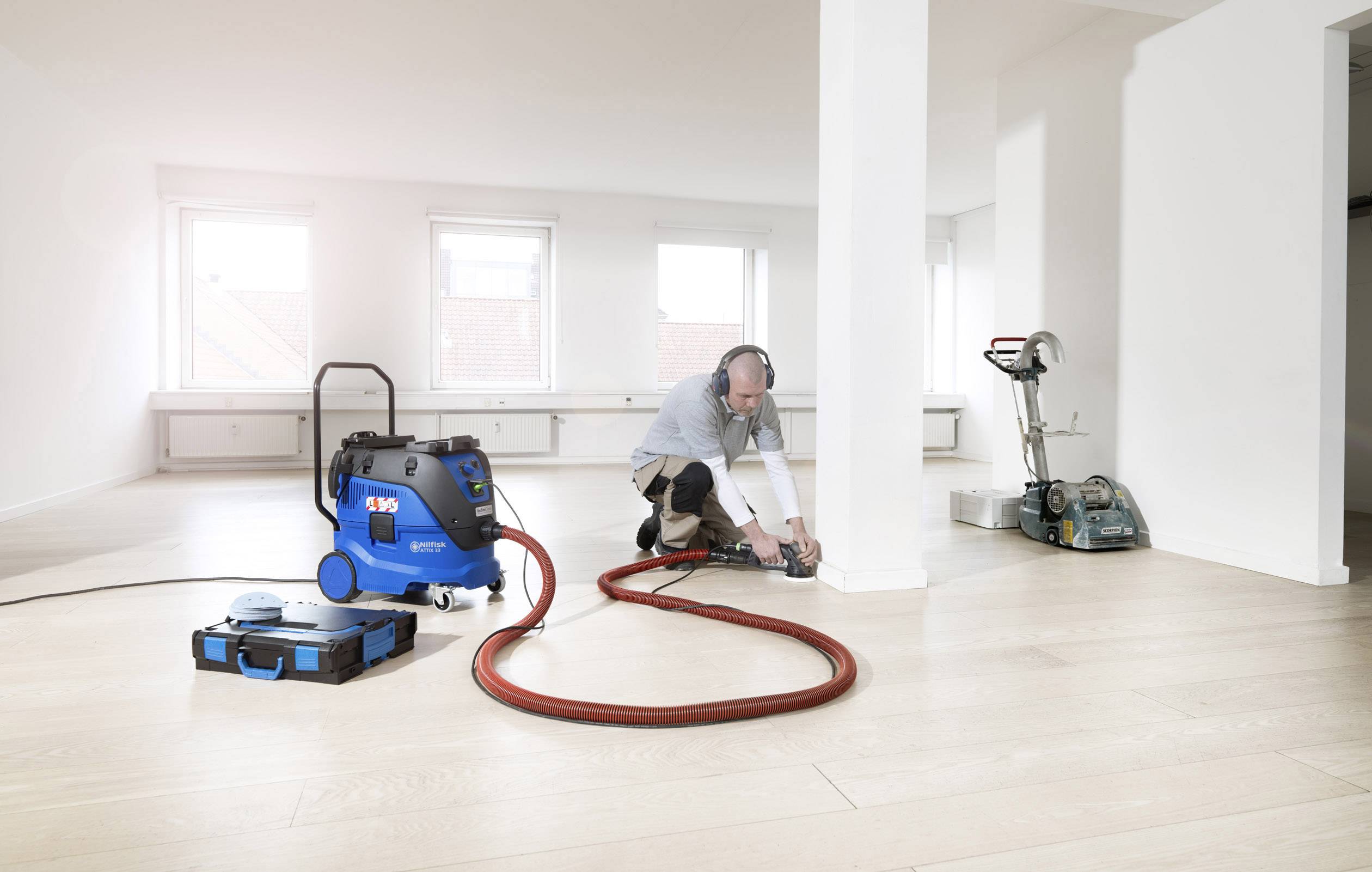 Een man bewerkt een houten vloer met een schuurapparaat in een lege ruimte, naast hem staat een stofzuiger.