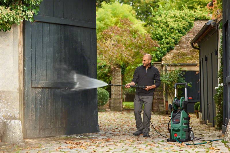 Een man reinigt een houten poort met een hogedrukreiniger in een tuin. Omgeven door bladeren en bomen bij daglicht.