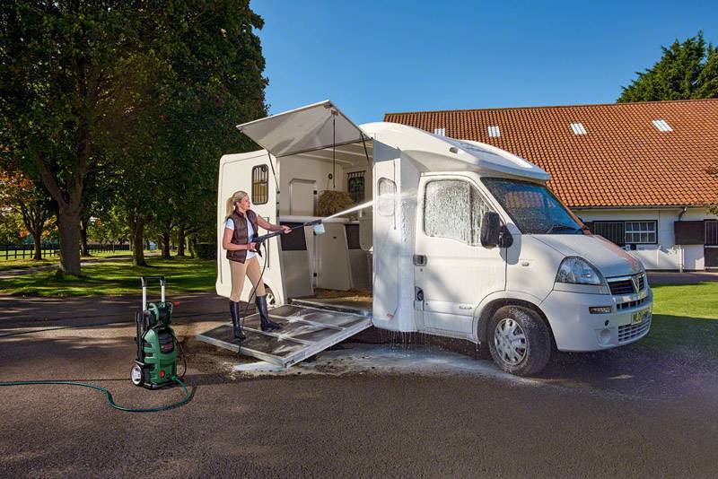 Een persoon reinigt een wit kampeerauto met een hogedrukreiniger op een zonnige parkeerplaats voor een gebouw met een rood dak.