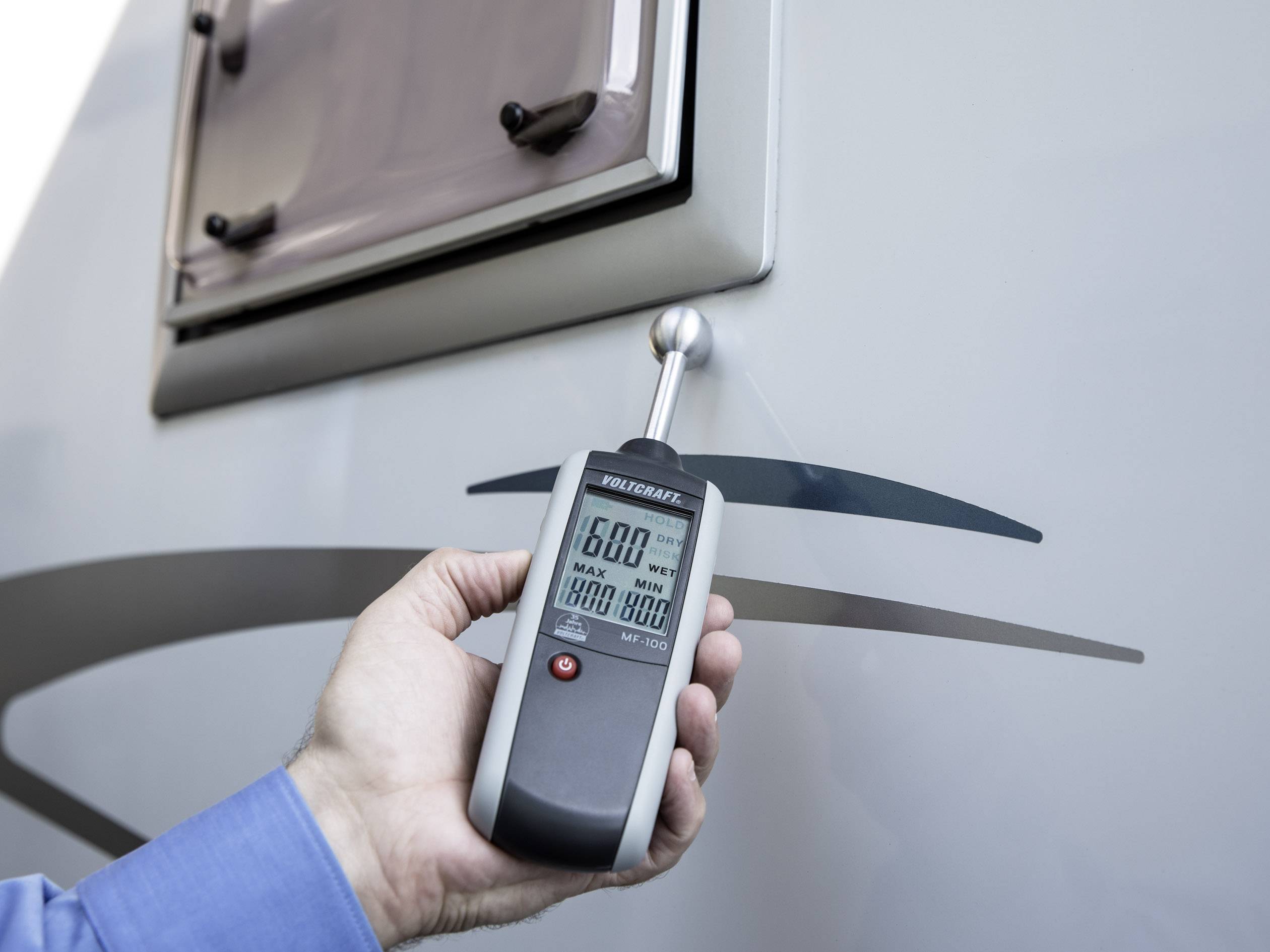 Een persoon houdt een vochtmeter tegen de buitenwand van een camper. Het display toont vochtigheidswaardes.