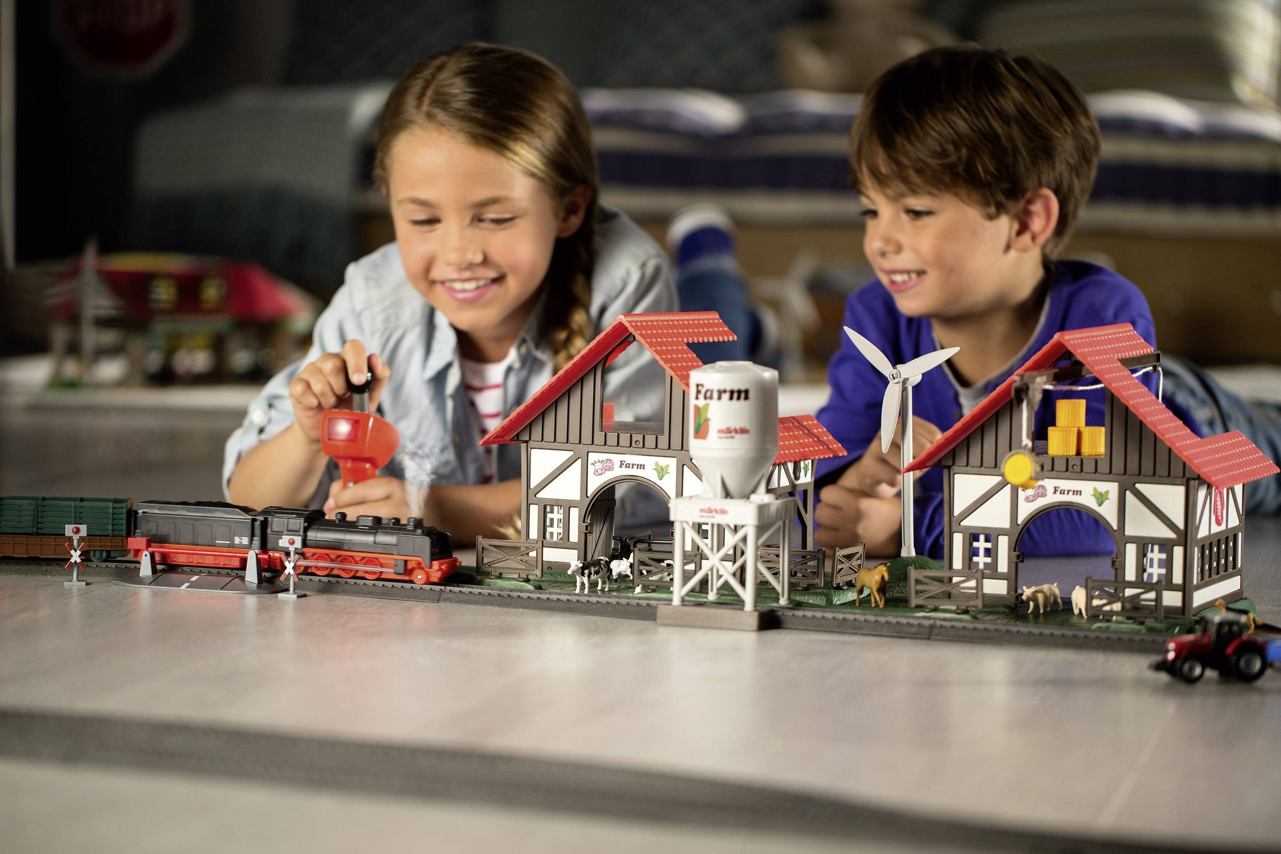 Twee kinderen spelen op de grond met een modelspoorbaan en gebouwen. De scène toont een idyllisch landschap met een boerderijthema.