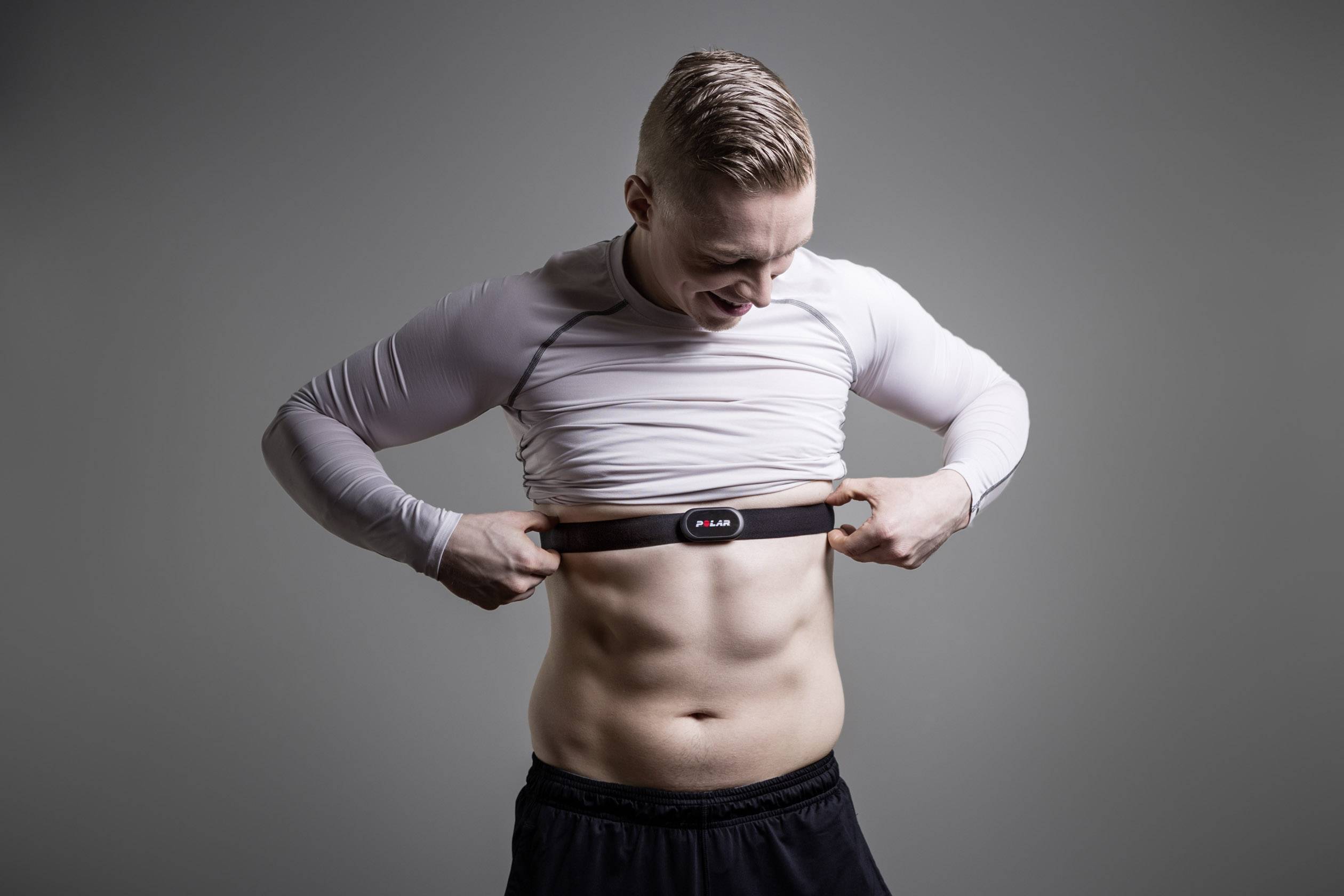 Een man in sportkleding bevestigt een hartslagmeter om zijn borst, glimlachend, voor een grijze achtergrond.