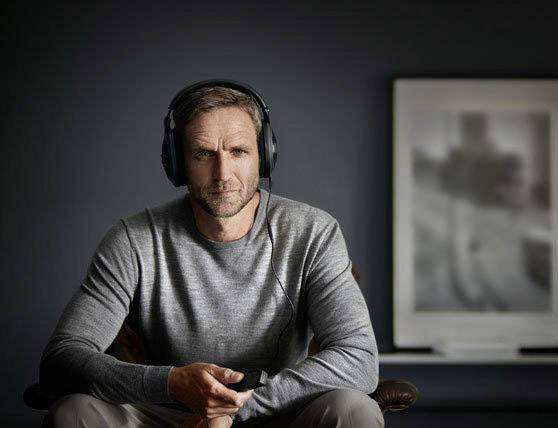 Een man van middelbare leeftijd met koptelefoon zit ontspannen en houdt een smartphone vast. De achtergrond is wazig met een groot beeld.