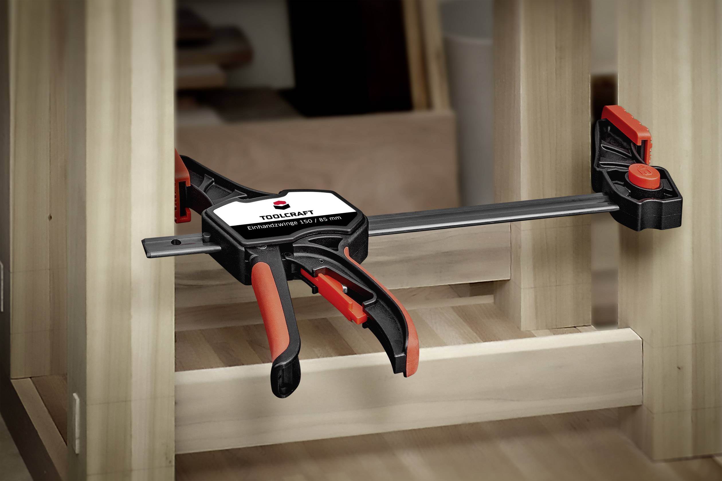 Een klem houdt twee houten onderdelen stevig bij elkaar. De klem is zwart met rode handgrepen en wordt gebruikt bij een meubelstuk.