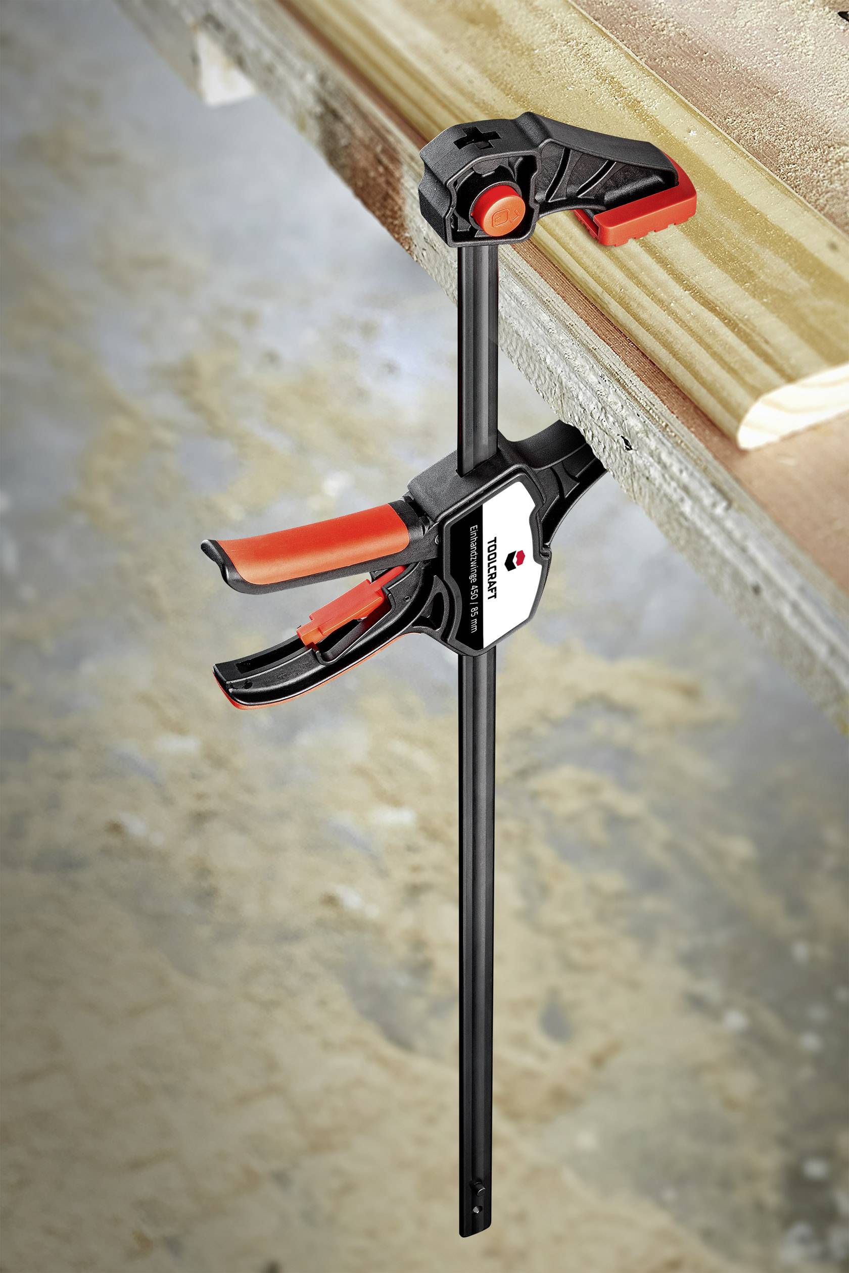 Een lijmklem houdt een houten plank vast op een werkbank. De lijmklem is zwart en rood met een ergonomische greep voor gemakkelijk klemmen.