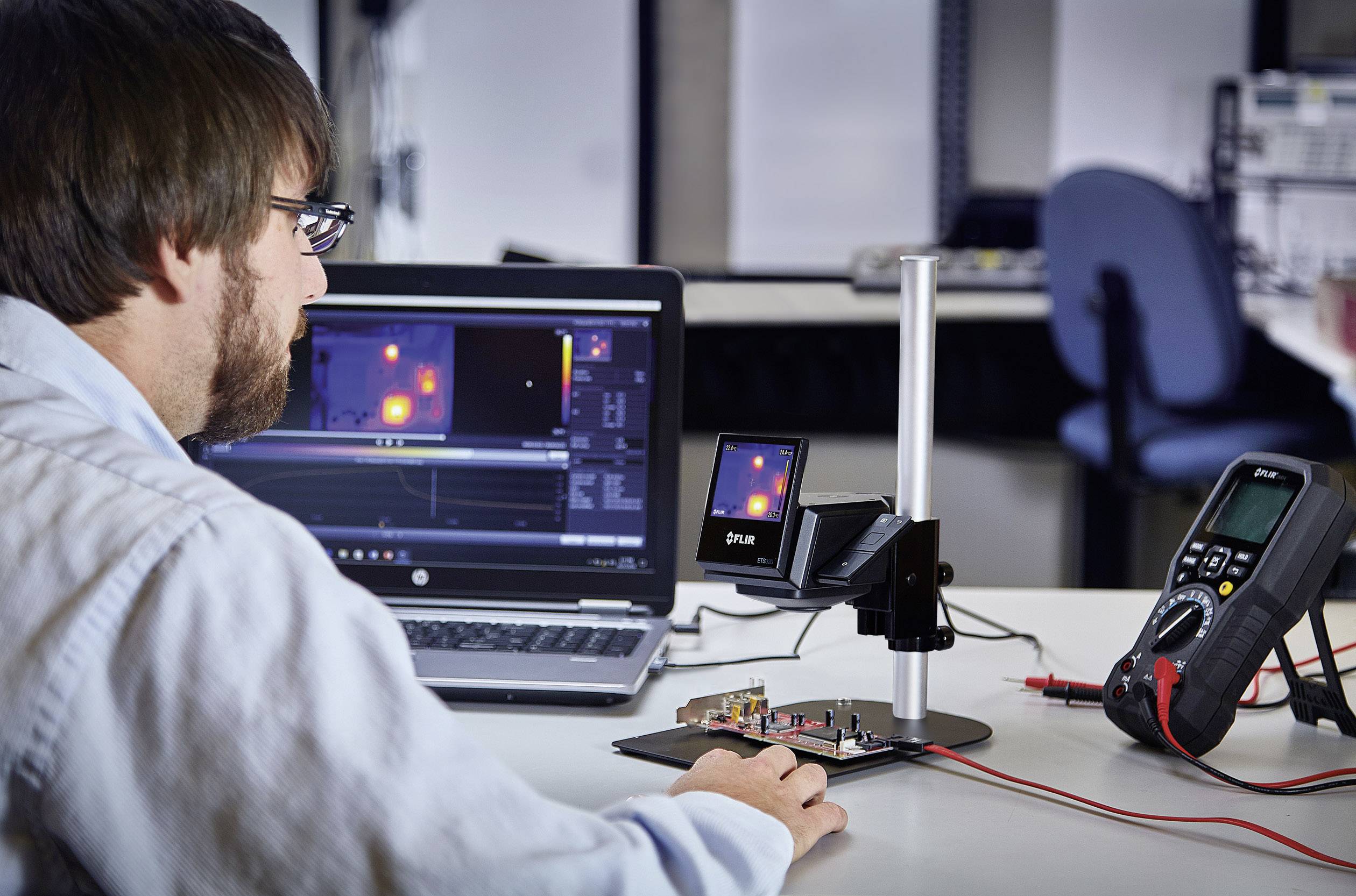 Ingenieur die gegevens analyseert op een laptop, met laborapparatuur en warmtebeeldcamera op een tafel. Focus op elektronische testapparatuur.