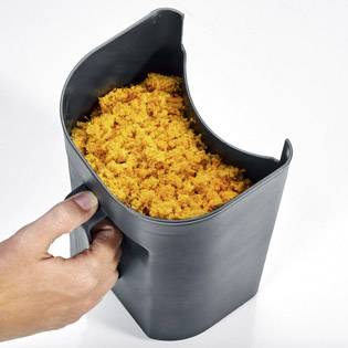 Een hand houdt een zwarte container vast, gevuld met oranje, verkruimelde wortel, vermoedelijk afval van een sapcentrifuge.