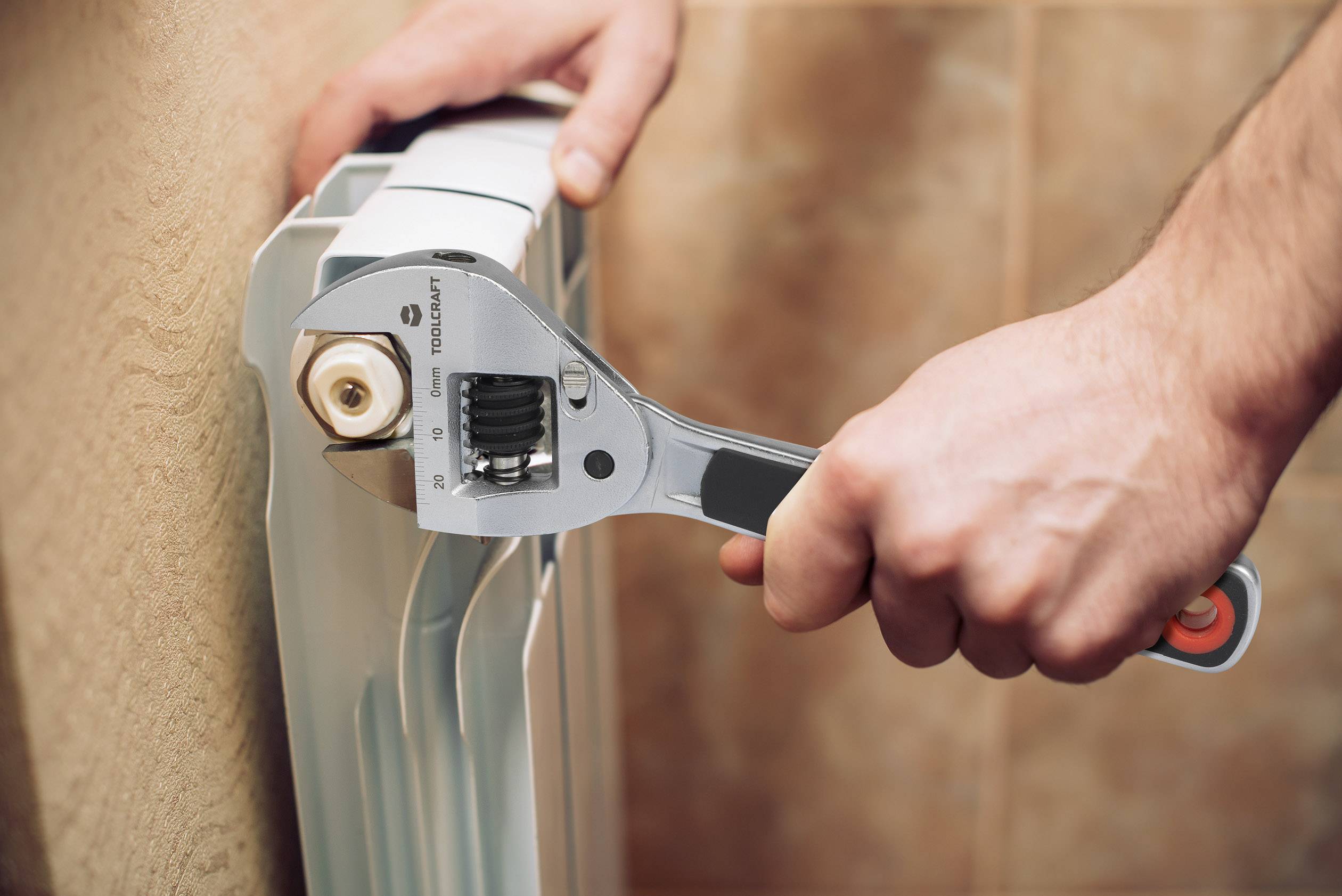 Een persoon draait met een verstelbare moersleutel de kop van een radiatorventiel vast aan een beige betegelde muur.