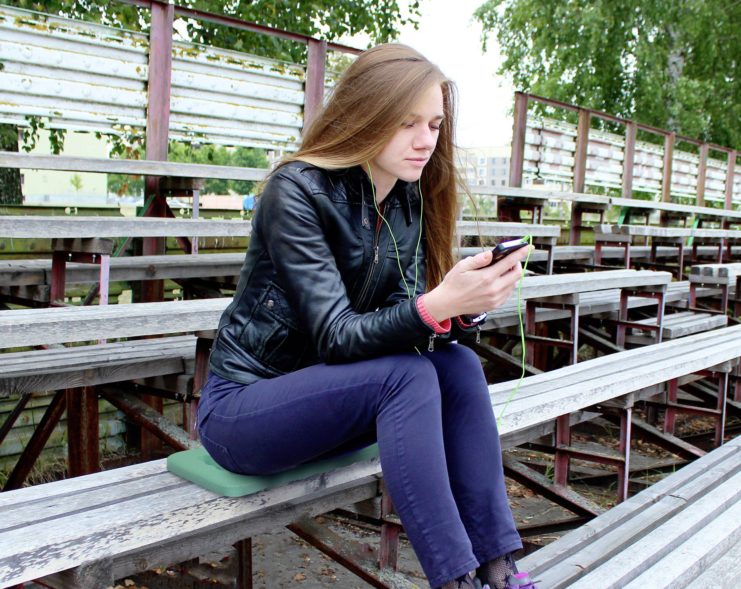 Een vrouw zit op houten banken buiten en kijkt naar haar mobiele telefoon. Ze draagt een zwarte leren jas en donkere broeken. De achtergrond toont bomen.