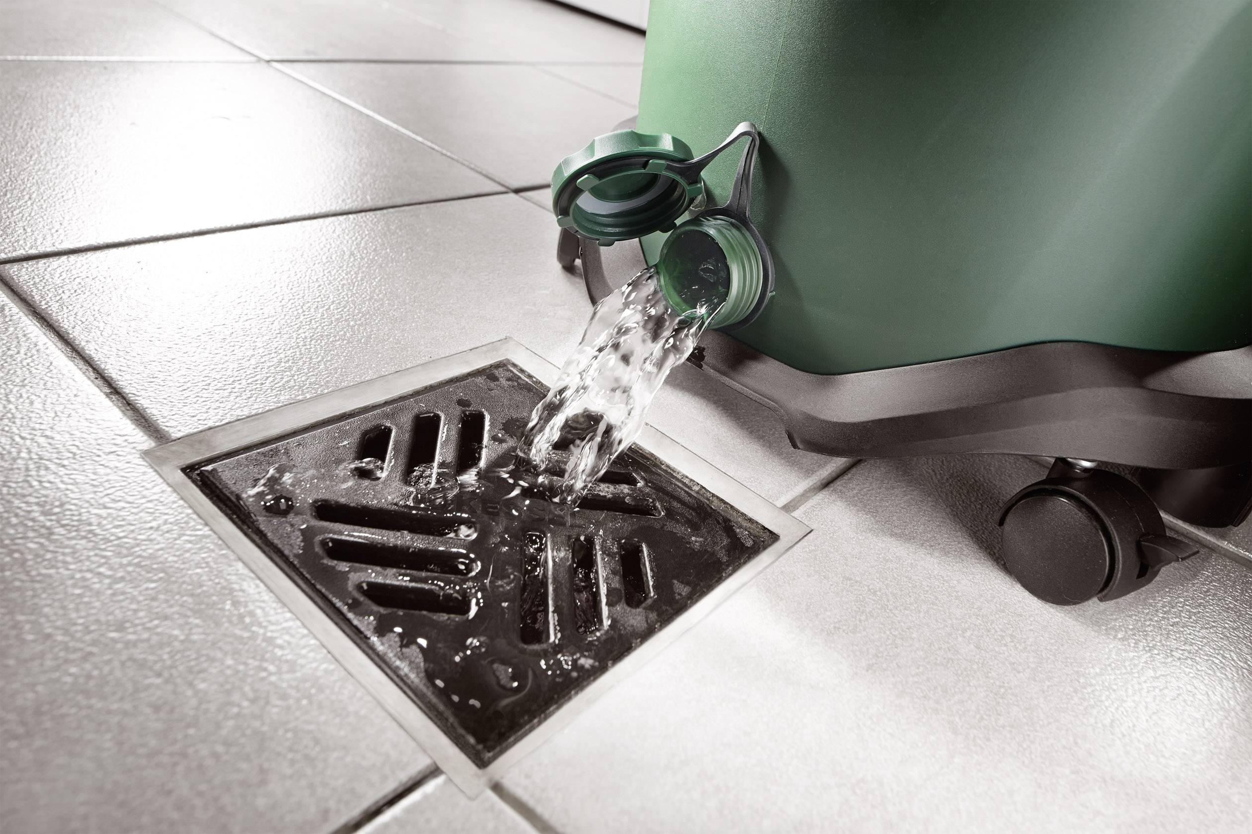 Een groene container lekt water door een afvoerdeksel op een betegelde vloer.