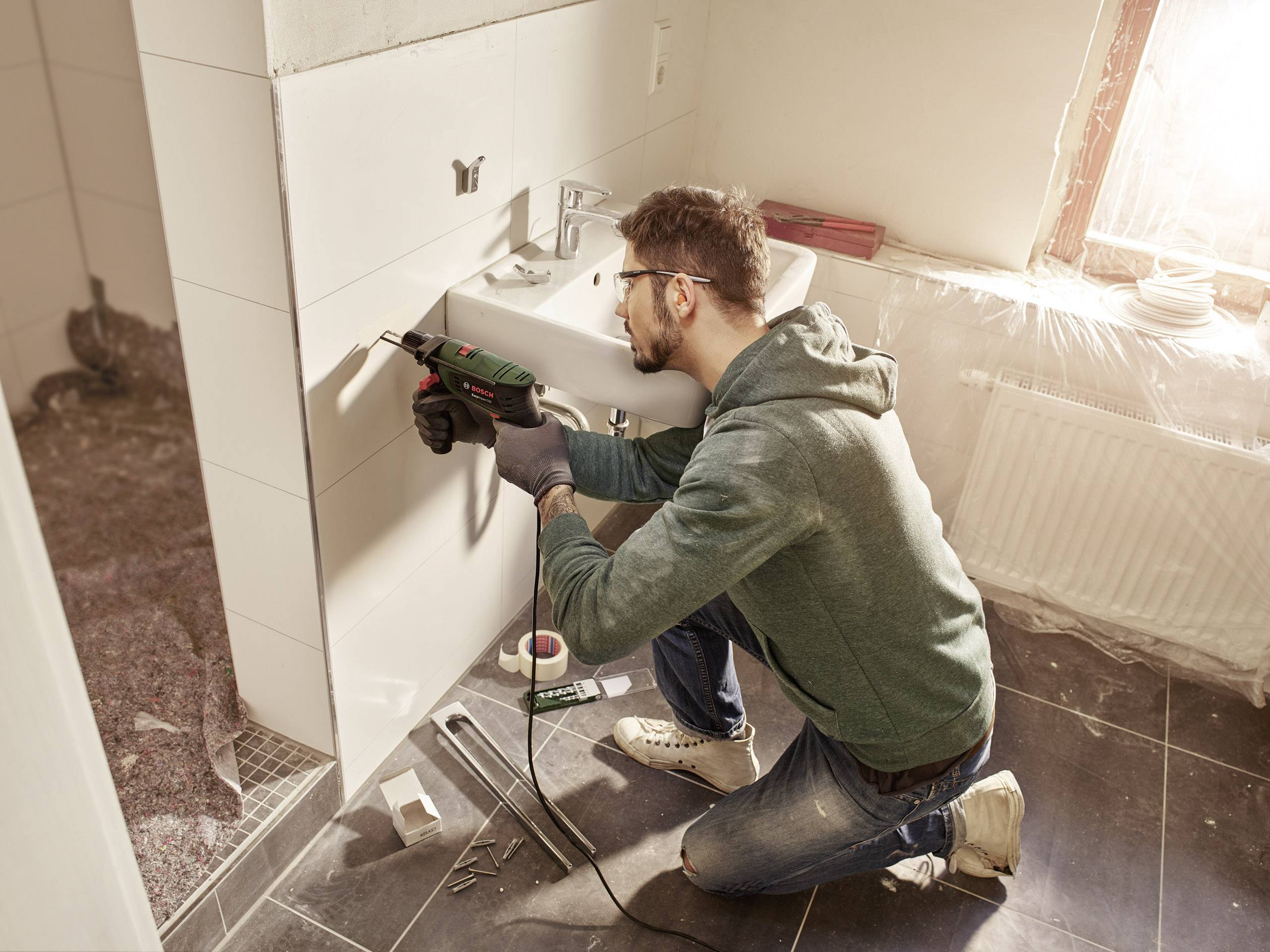 Een man in een groene hoodie boort een gat in een witte badkamermuur naast een wastafel, omringd door renovatiegereedschap.