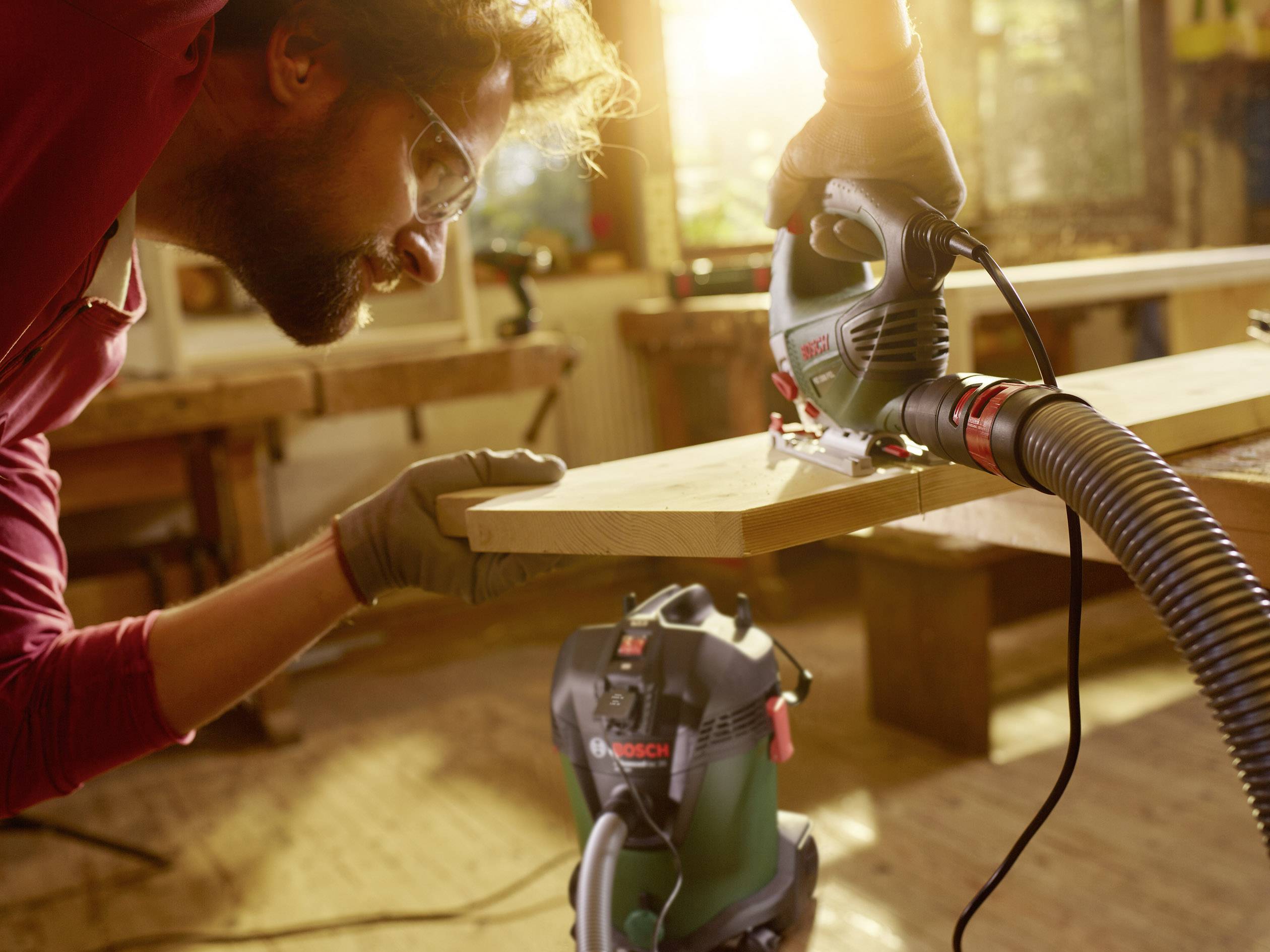 Een man werkt geconcentreerd met een elektrische decoupeerzaag aan een stuk hout in een werkplaats. Zonlicht valt door het raam.