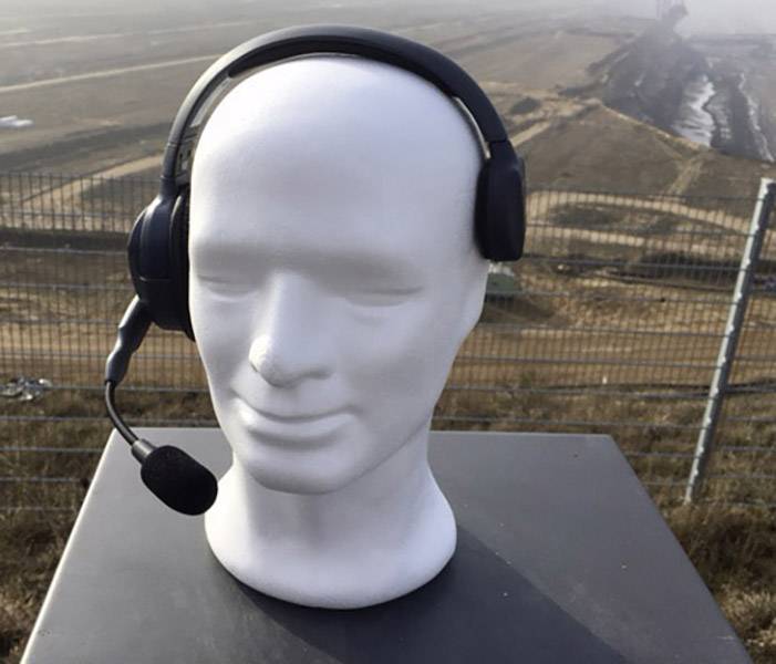 Een witte etalagepop met een zwarte headset staat buiten voor een uitgestrekt, mistig landschapsachtergrond.