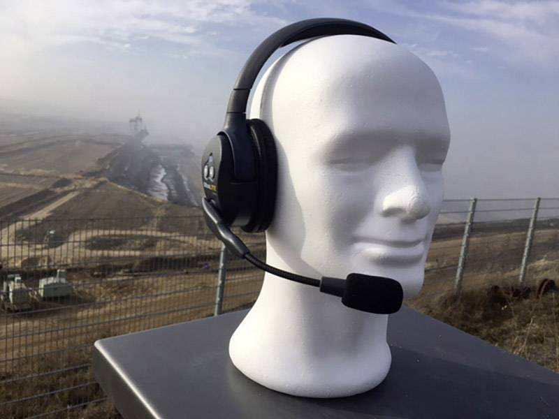 Een wit hoofdmodel met een zwarte headset staat buiten; op de achtergrond is een industrieel landschap zichtbaar.