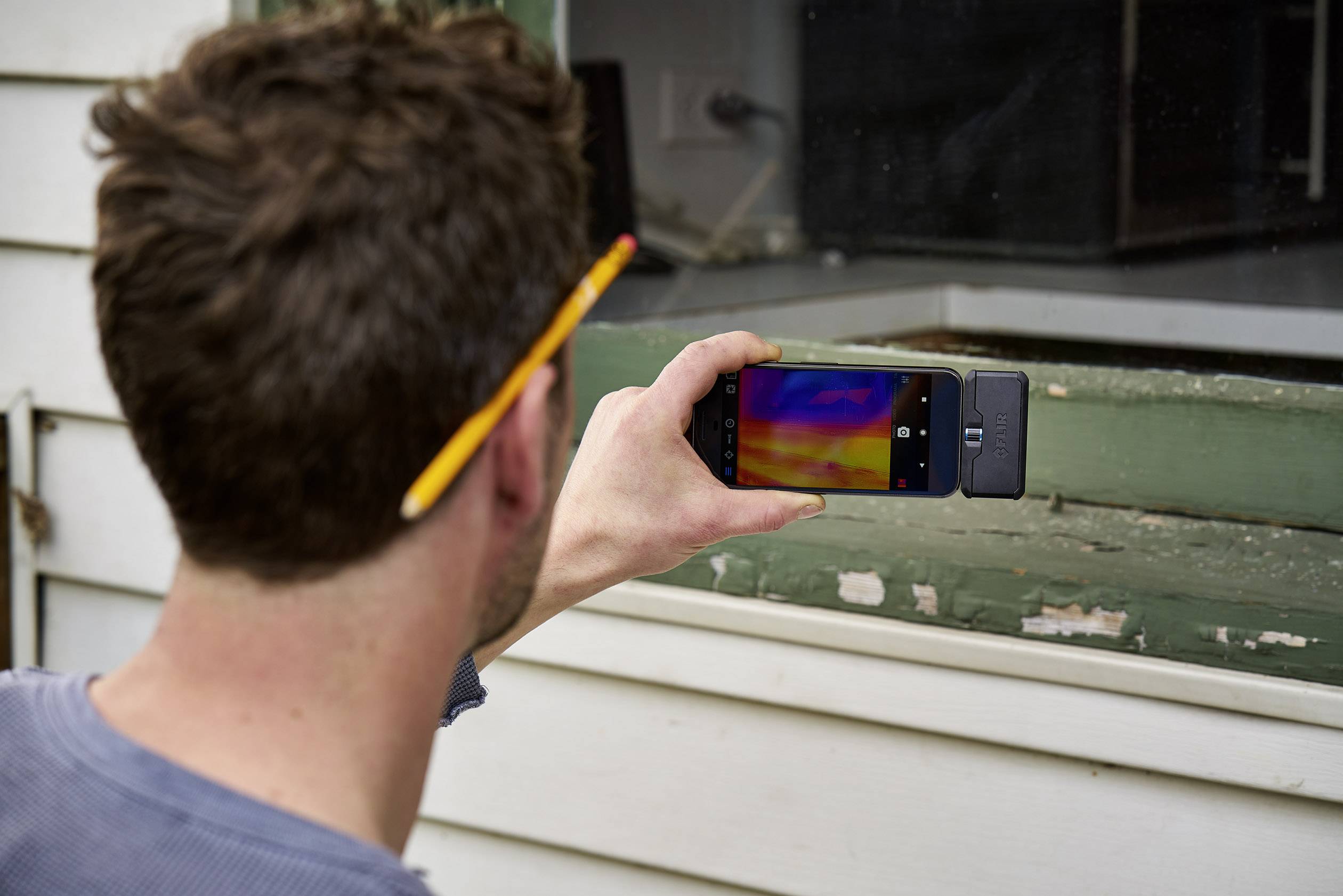 Een persoon houdt een smartphone met warmtebeeldcamera voor een raam om temperatuurverschillen in de gevel te analyseren.
