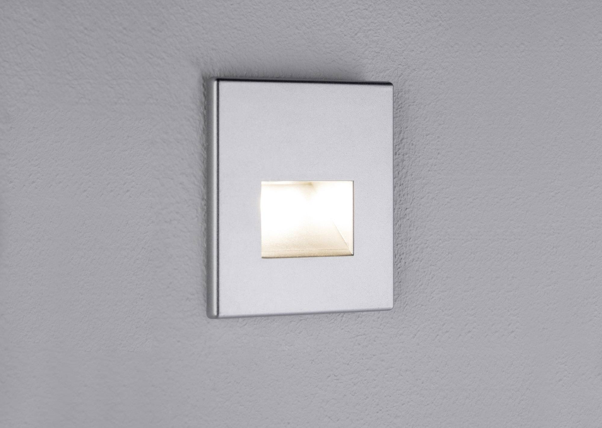 Een hoekige, zilveren wandlamp verspreidt zacht licht op een grijze muur, zonder verdere decoratieve elementen.
