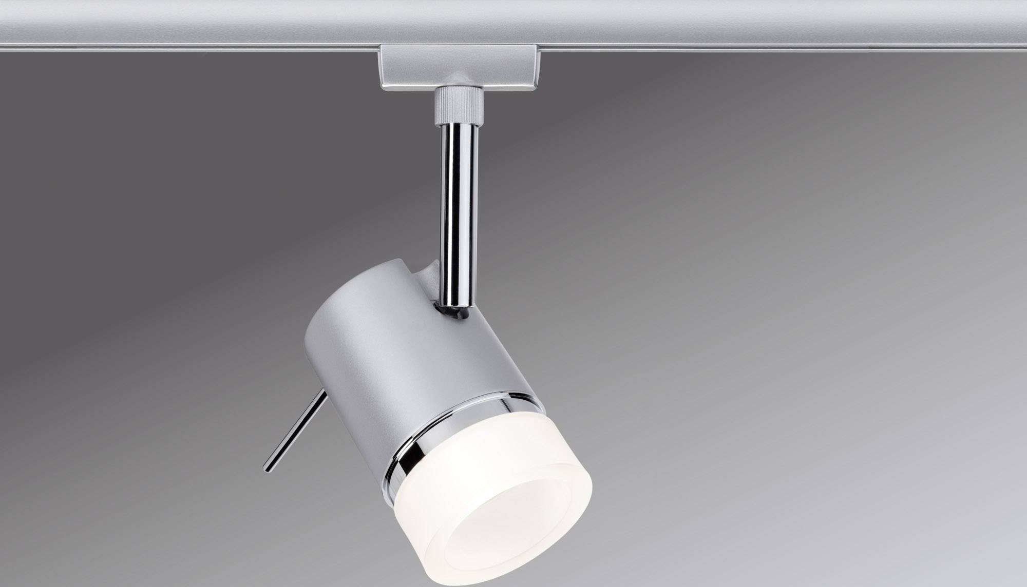 Een zilveren plafondlamp met een cilindrische lichtbron is gemonteerd op een rail en schijnt op een gladde, grijze oppervlakte.