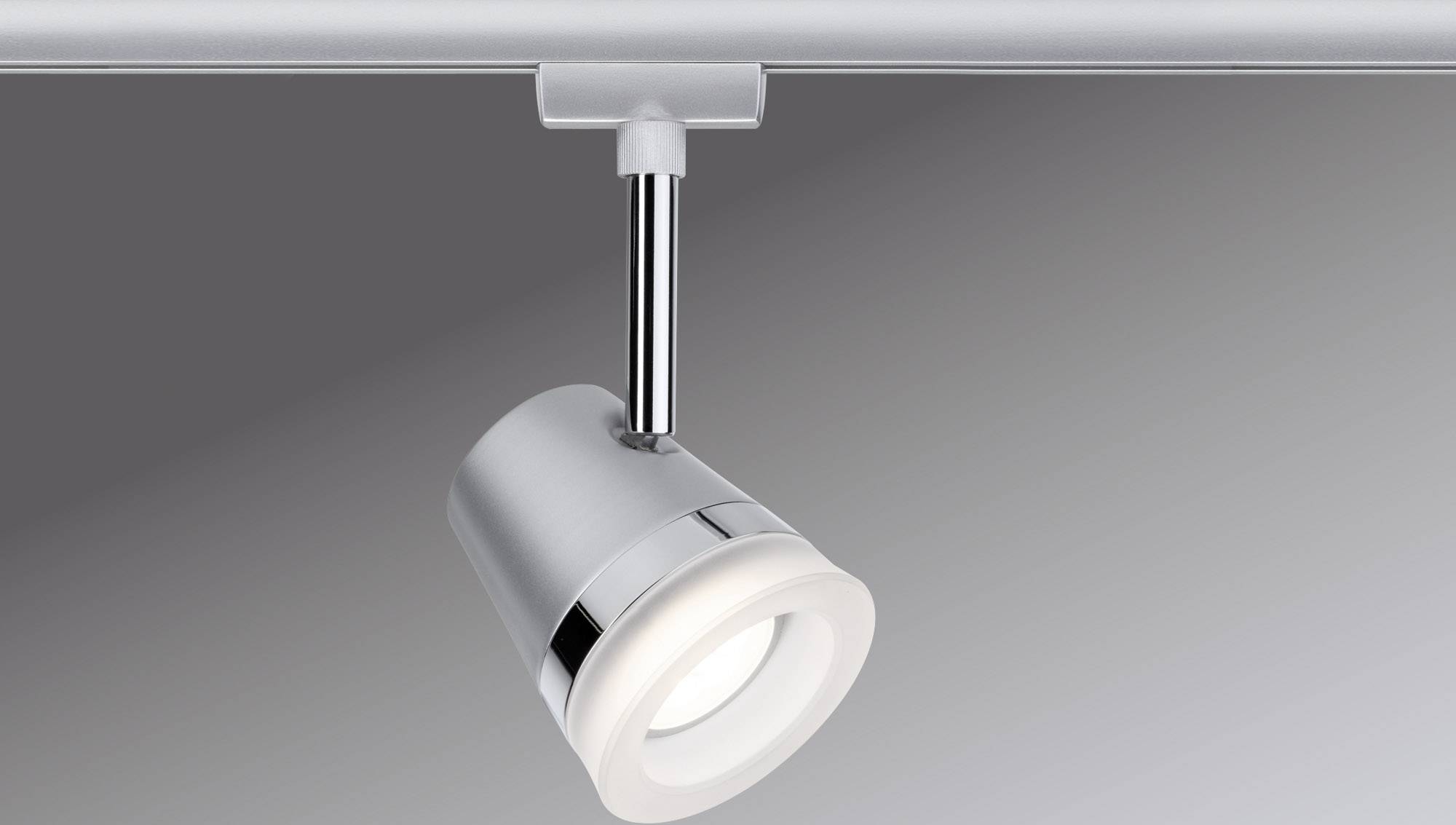 Een zilverkleurige plafondspot is gemonteerd aan een rail, verlicht. De achtergrond is grijs.