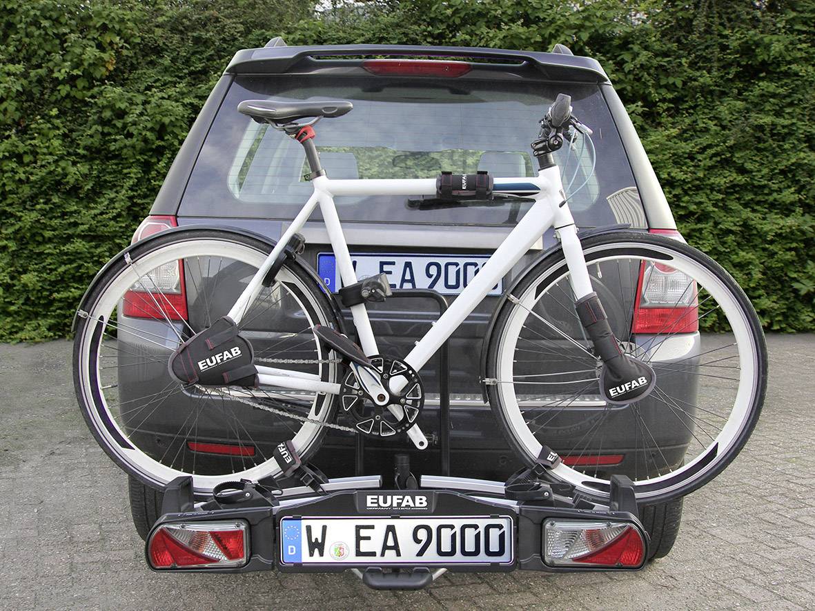 Een witte fiets is bevestigd op een fietsendrager aan de achterkant van een zwarte auto, die op een geplaveid parkeerterrein staat.