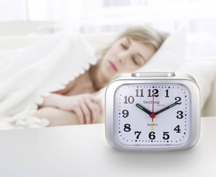 Een vrouw slaapt onder een deken op de achtergrond. Op de voorgrond staat een wekker die 7:10 uur aangeeft.