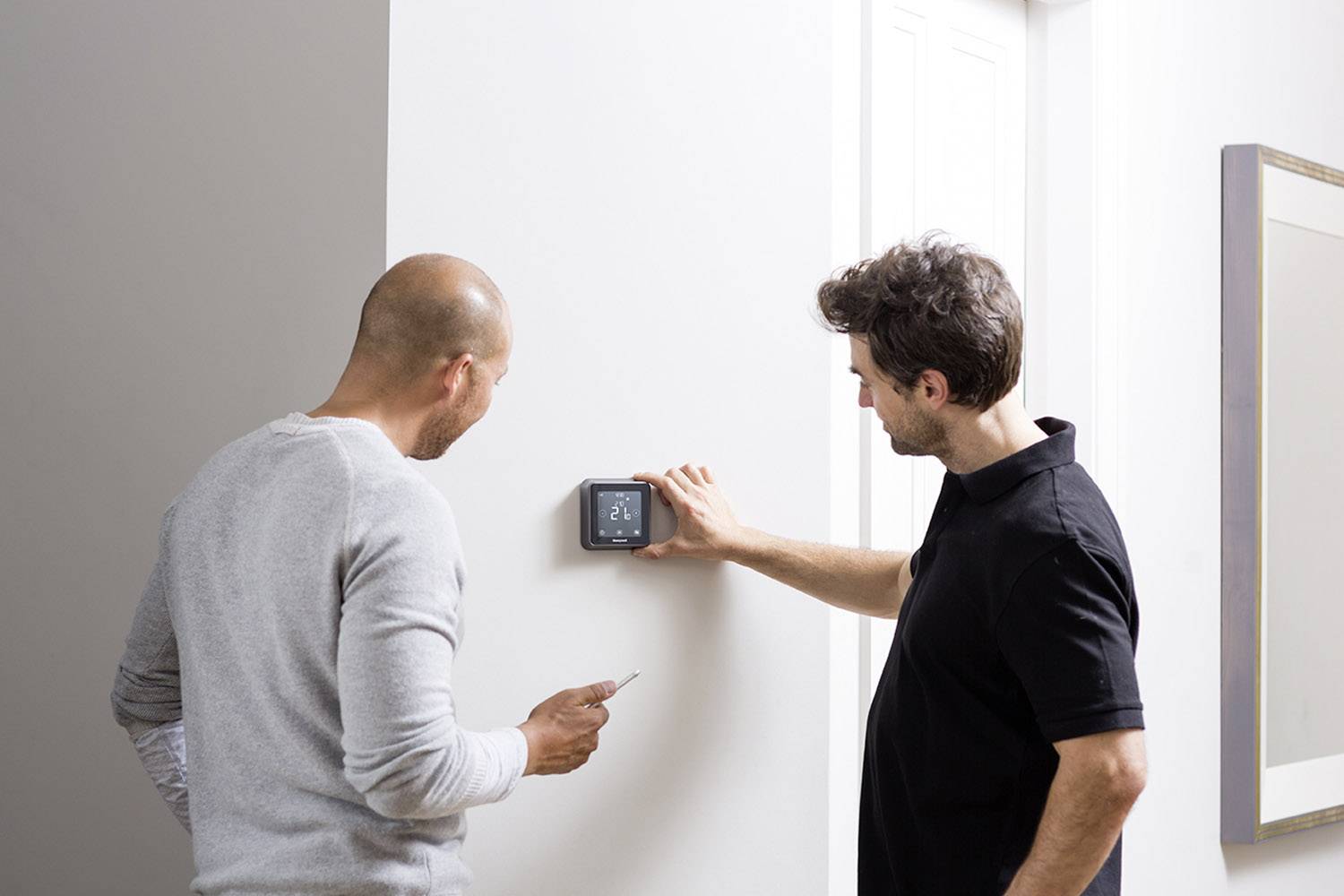 Twee mannen bij een witte muur, waarbij een een digitale thermostaat bedient en de ander een notitieboek vasthoudt.